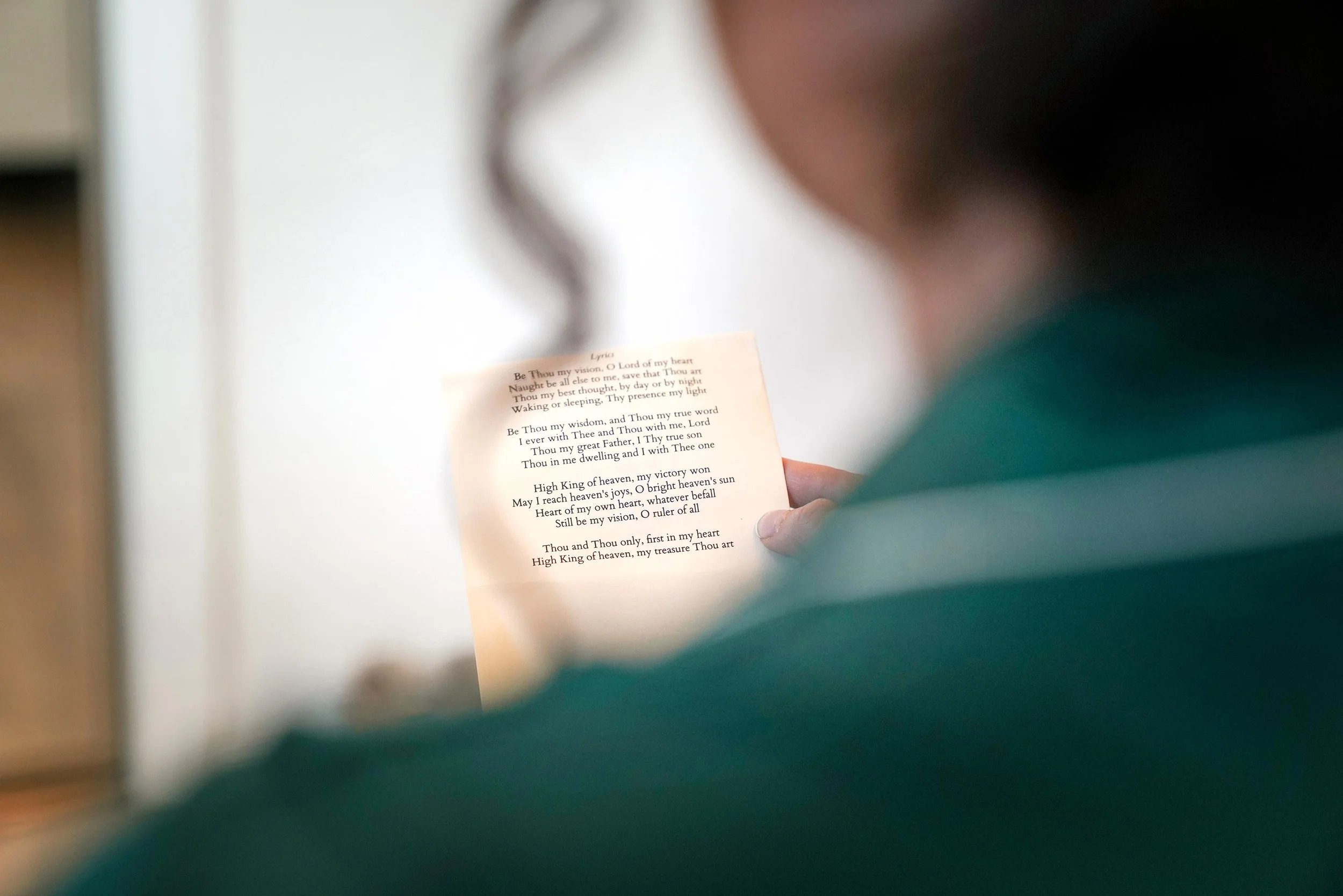 A close-up shot of the back of a bridesmaid wearing a green top, holding a paper with the lyrics to the hymn "Be Thou My Vision" during a wedding ceremony or rehearsal.



