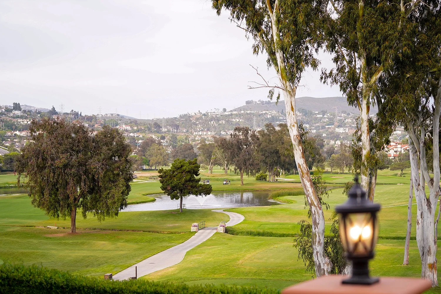 An elevated view of a lush green golf course landscape featuring a pond with a fountain, a paved cart path, tall eucalyptus trees, and rolling hills with residential homes in the background under an overcast sky.

