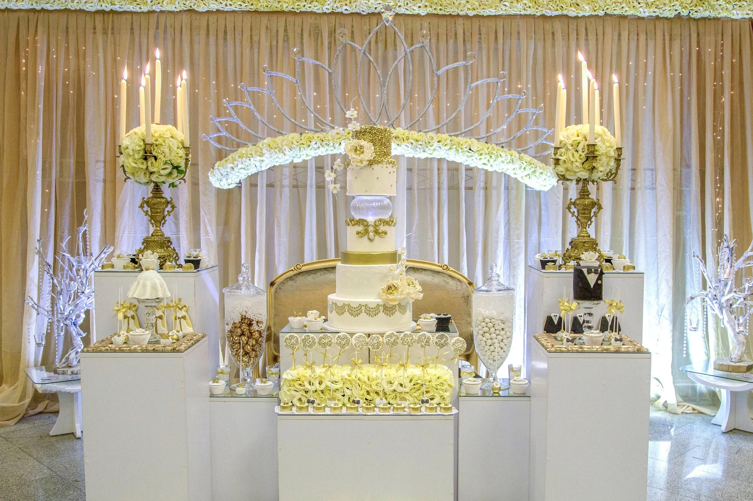An elaborate, formal wedding dessert table featuring a multi-tiered white cake with gold accents, flanked by white floral arrangements on gold stands with tall candles, assorted treats in clear jars, and decorative white display items.