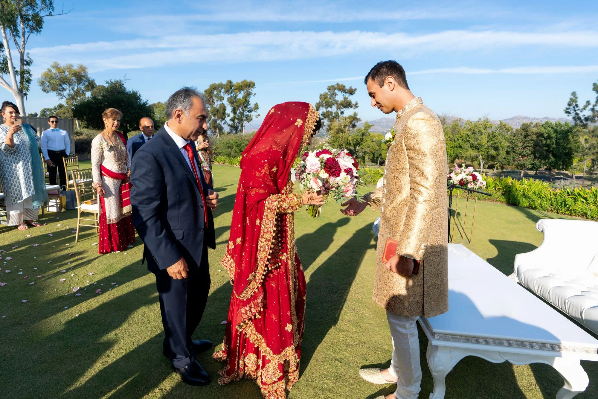 An outdoor photograph capturing a moment during a wedding ceremony or event. A bride in a traditional, ornate red lehenga and veil is facing the groom, who is wearing a gold brocade sherwani. An older man in a navy suit with a red tie stands to the l