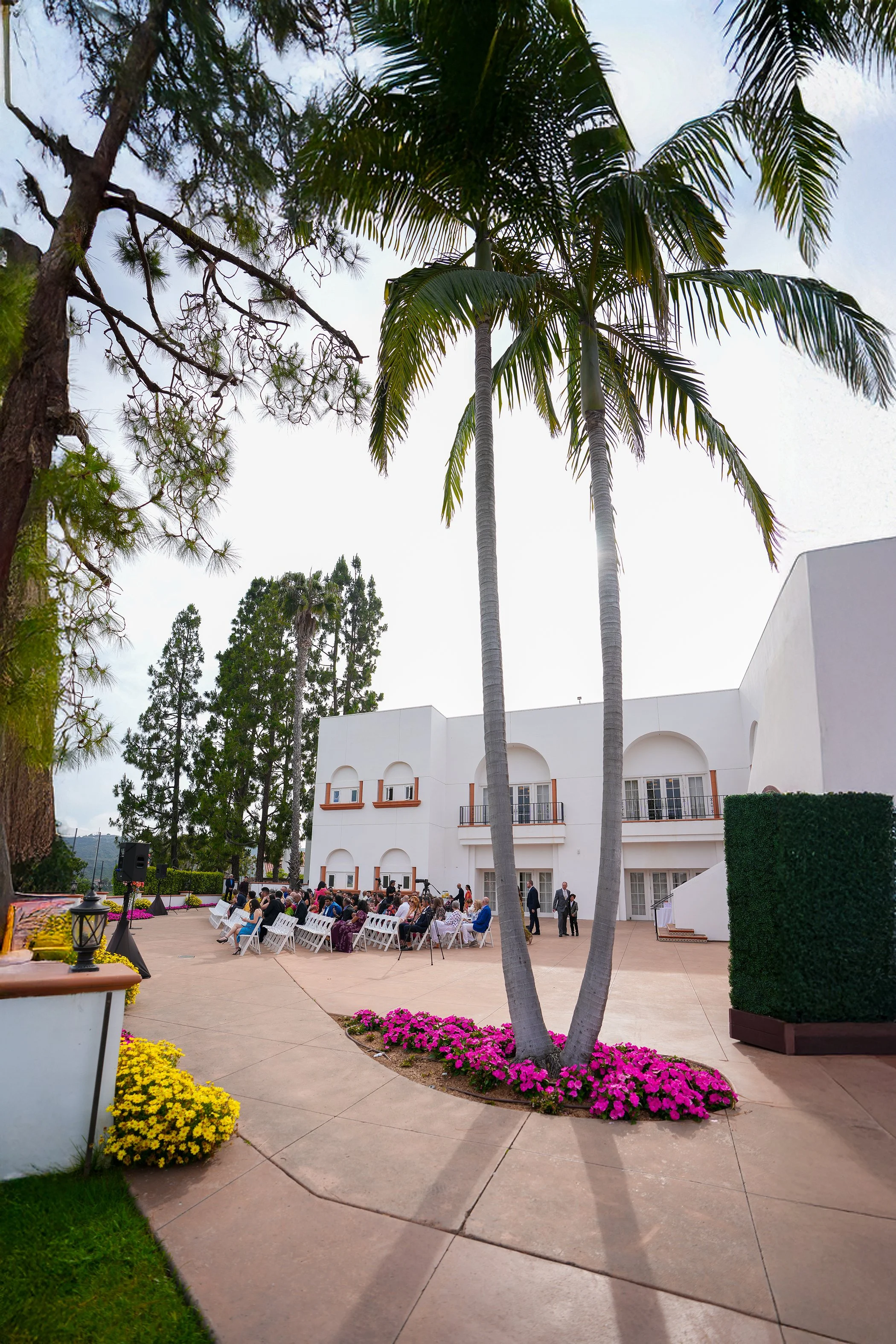 A wide-angle outdoor wedding ceremony shot by Reimo Photography at Omni resort in La Costa golf club. Guests are seated in white chairs on a paved patio, facing away from the camera toward a white two-story building with arched balconies. Tall palm t