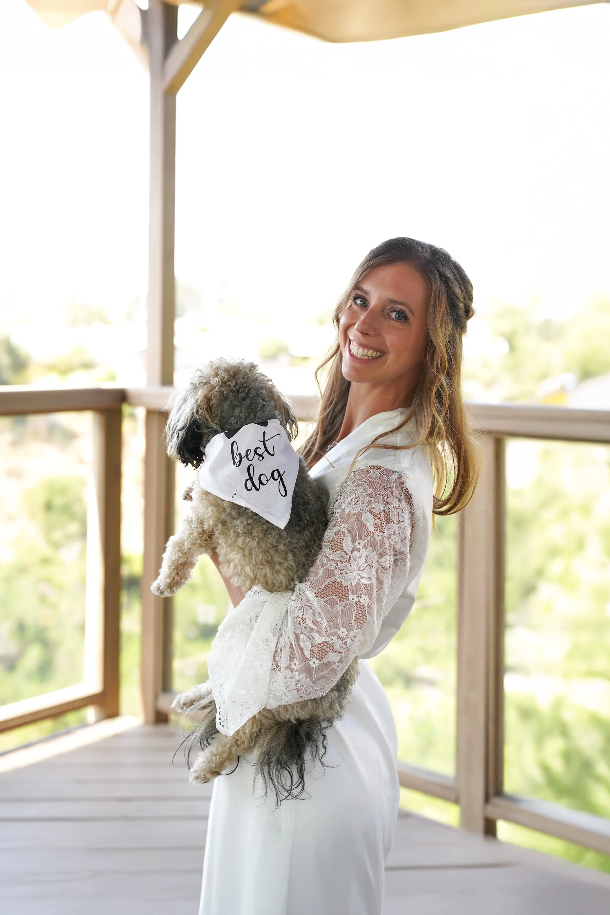 A smiling bride in a white lace robe holding her small gray dog wearing a 'Best Dog' bandana on a balcony.