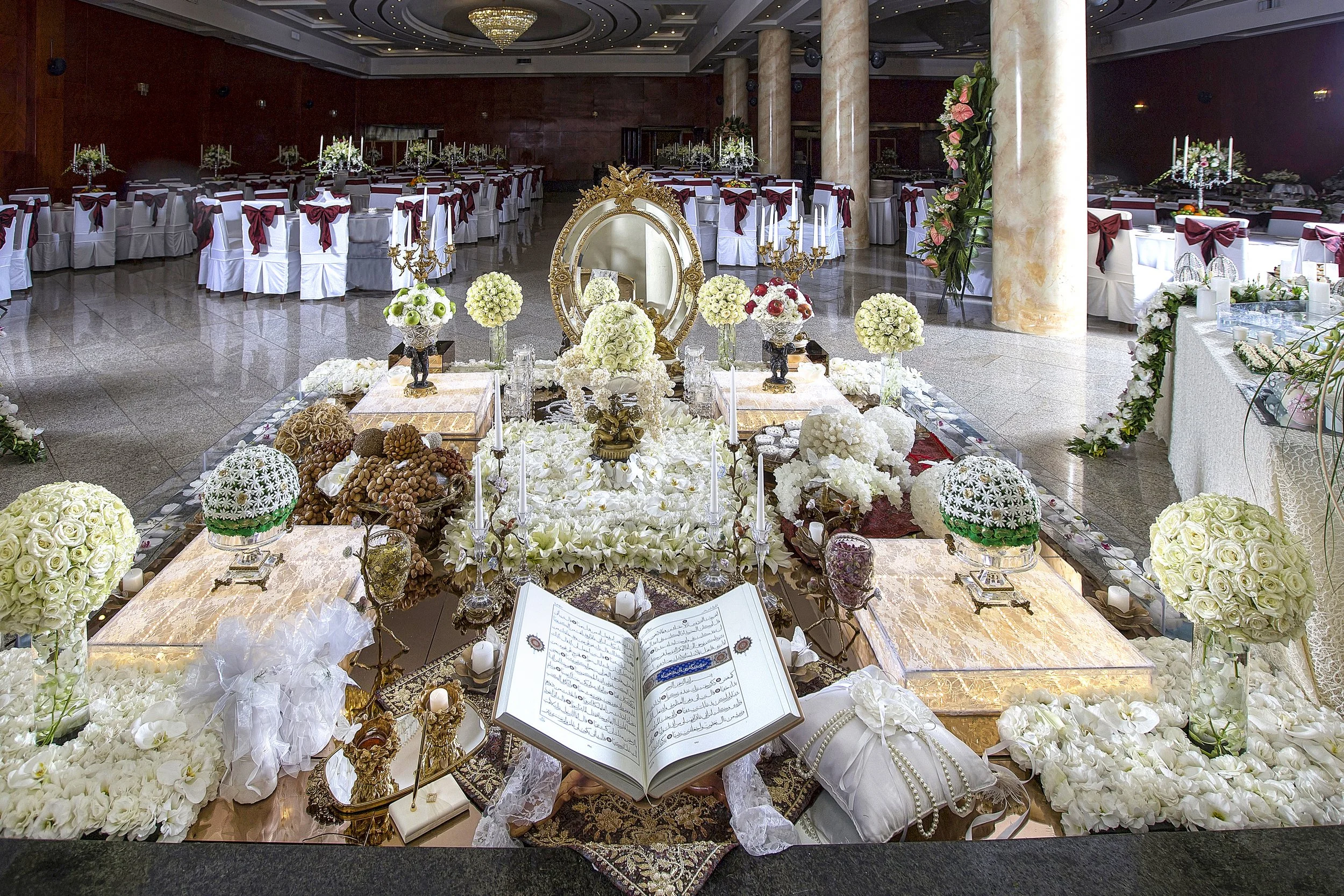 An elaborate, traditional Persian wedding table setting (Sofreh Aghd) featuring an ornate gold mirror, candelabras, a holy book, nuts, sweets, and flowers on a white lace tablecloth.