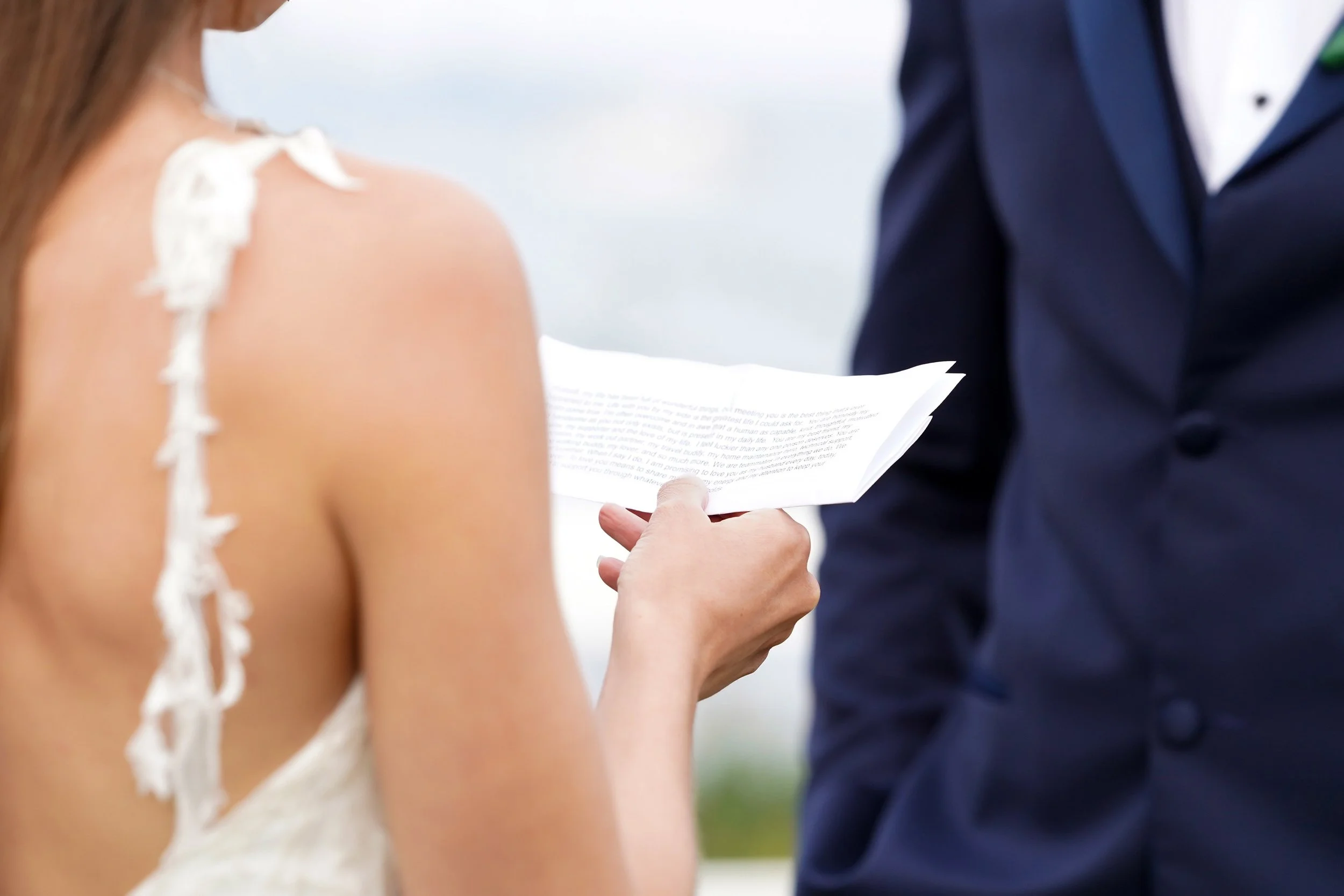 A close-up, over-the-shoulder shot of a bride in a white lace dress holding a piece of paper and reading aloud, standing next to a groom in a navy blue suit.