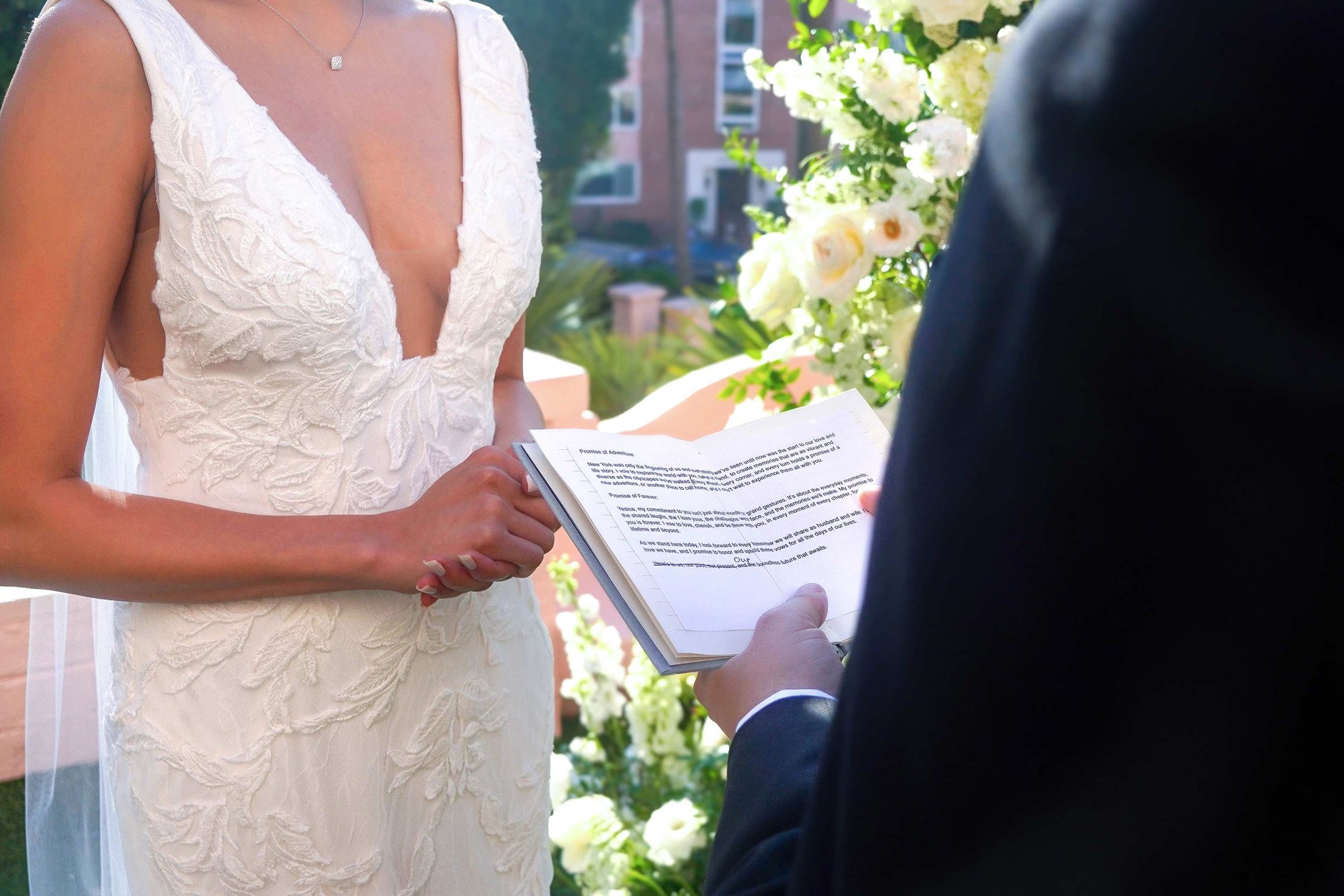 A close-up photograph capturing a bride and groom exchanging vows during an outdoor wedding ceremony. The bride, wearing a white lace dress and a delicate necklace, holds a small booklet of vows, facing the groom, whose back is to the camera in a dar
