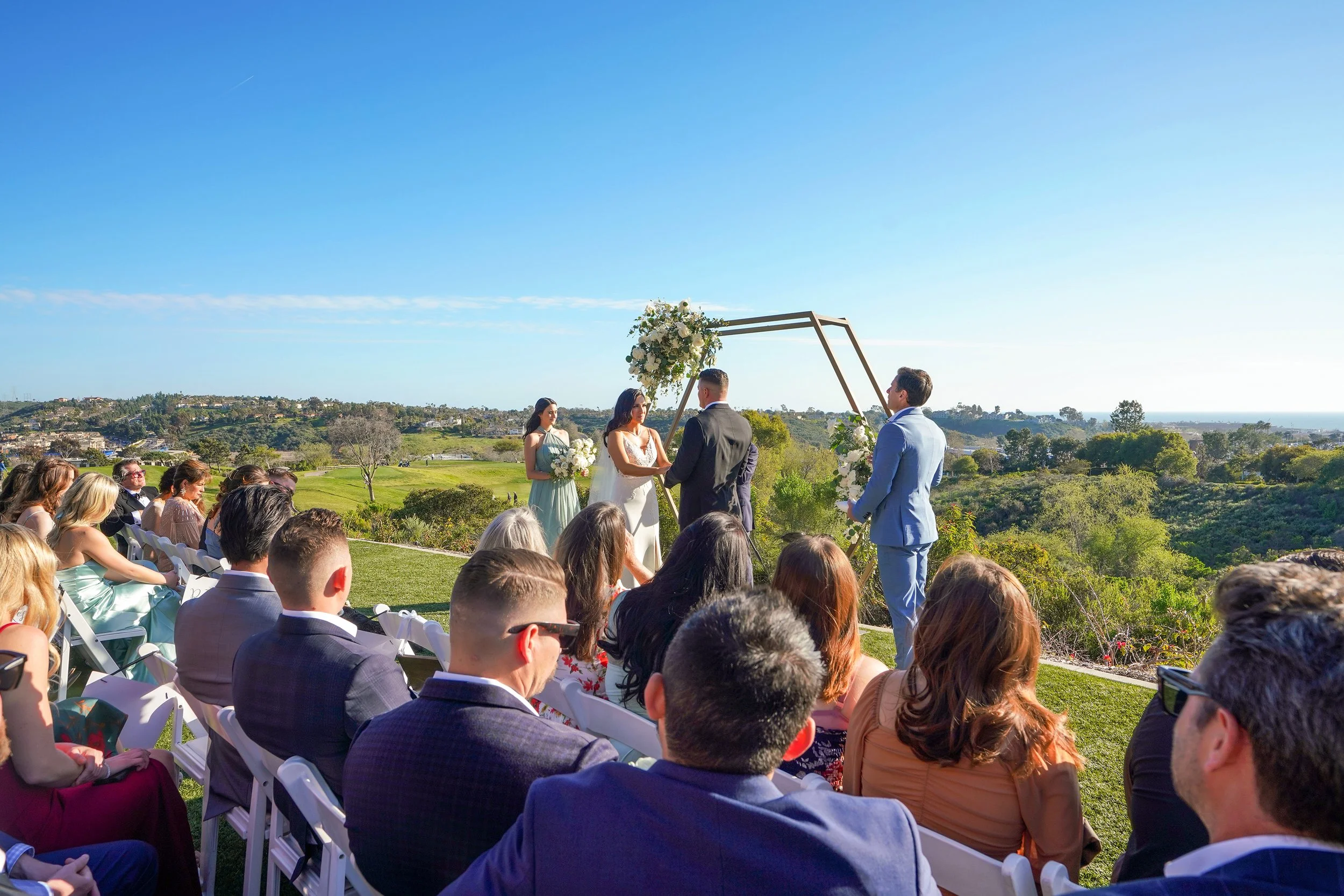 An outdoor wedding ceremony is in progress under a clear blue sky. The bride wears a white, floor-length, bias-cut slip gown with spaghetti straps. Beside her, the groom is dressed in a dark, classic-fit charcoal or black suit. An officiant in a ligh