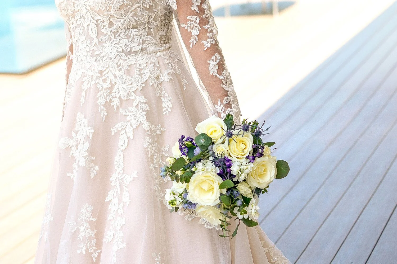 A bride wearing a long-sleeved, lace wedding dress holds a bouquet of white roses, purple wildflowers, and green foliage while standing on a light-colored wooden deck.