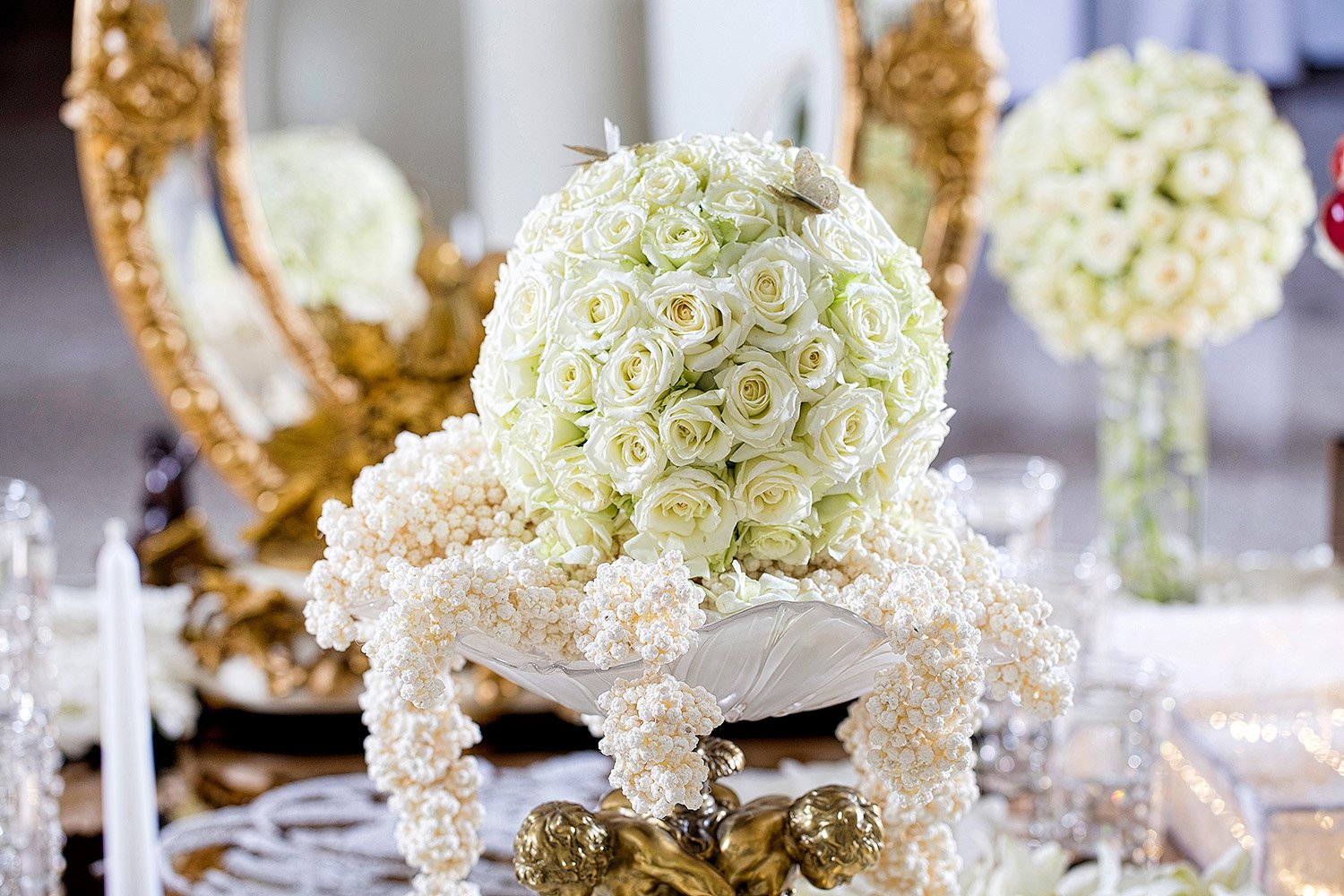 An ornate floral centerpiece featuring a sphere of white roses and draped white flower garlands, displayed on a gold stand with a large gold mirror and another floral arrangement in the background.