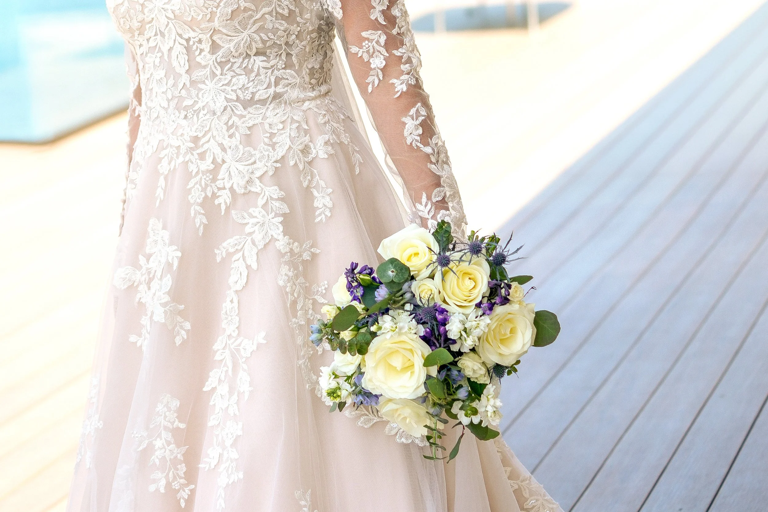 A bride in a long-sleeved lace wedding dress holds a bouquet of yellow and white roses, purple thistles, and greenery.