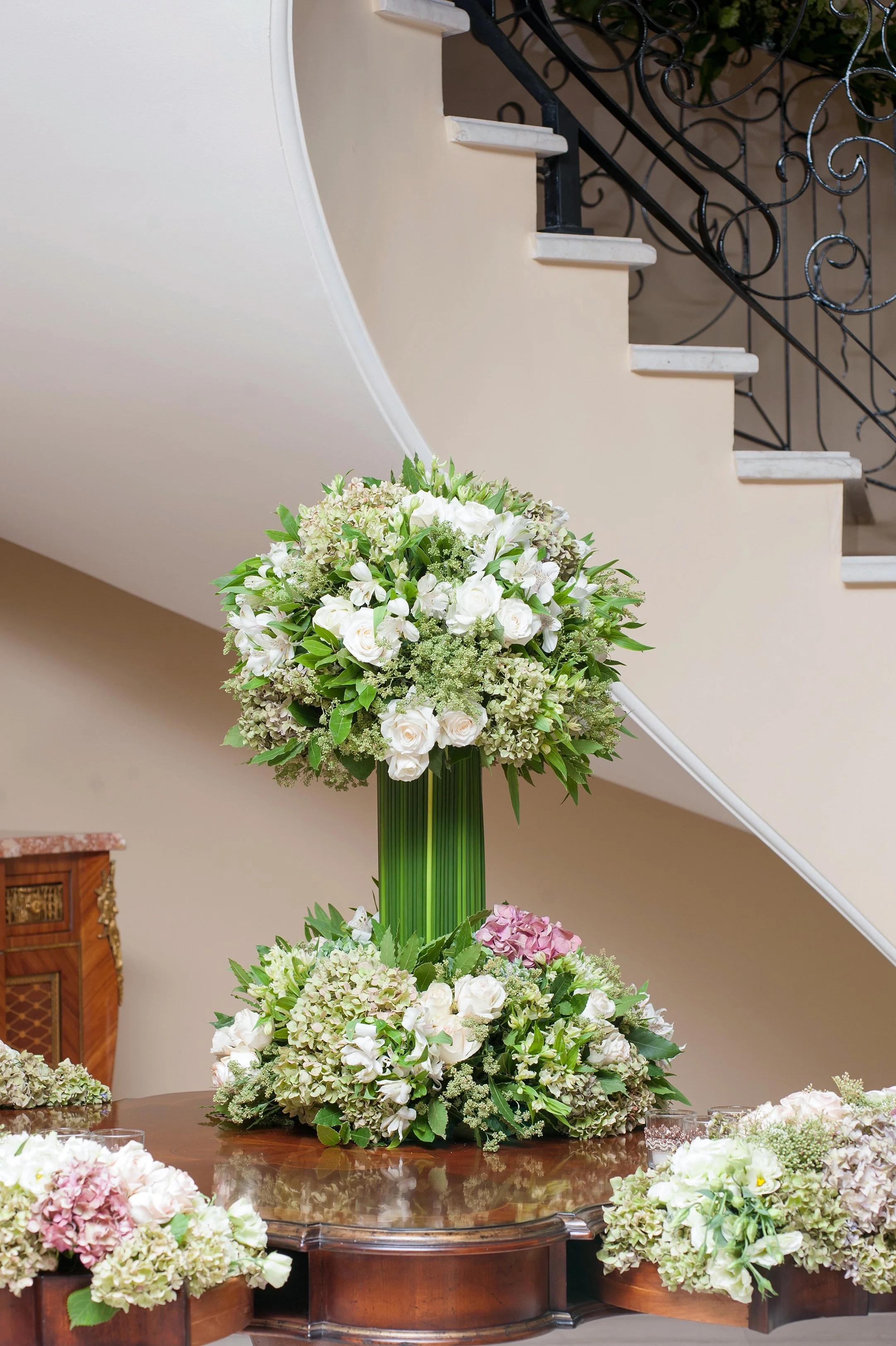 A large, elegant white and green floral arrangement in a tall glass vase on a wooden table in an entryway, with a curved staircase and wrought-iron railing in the background.