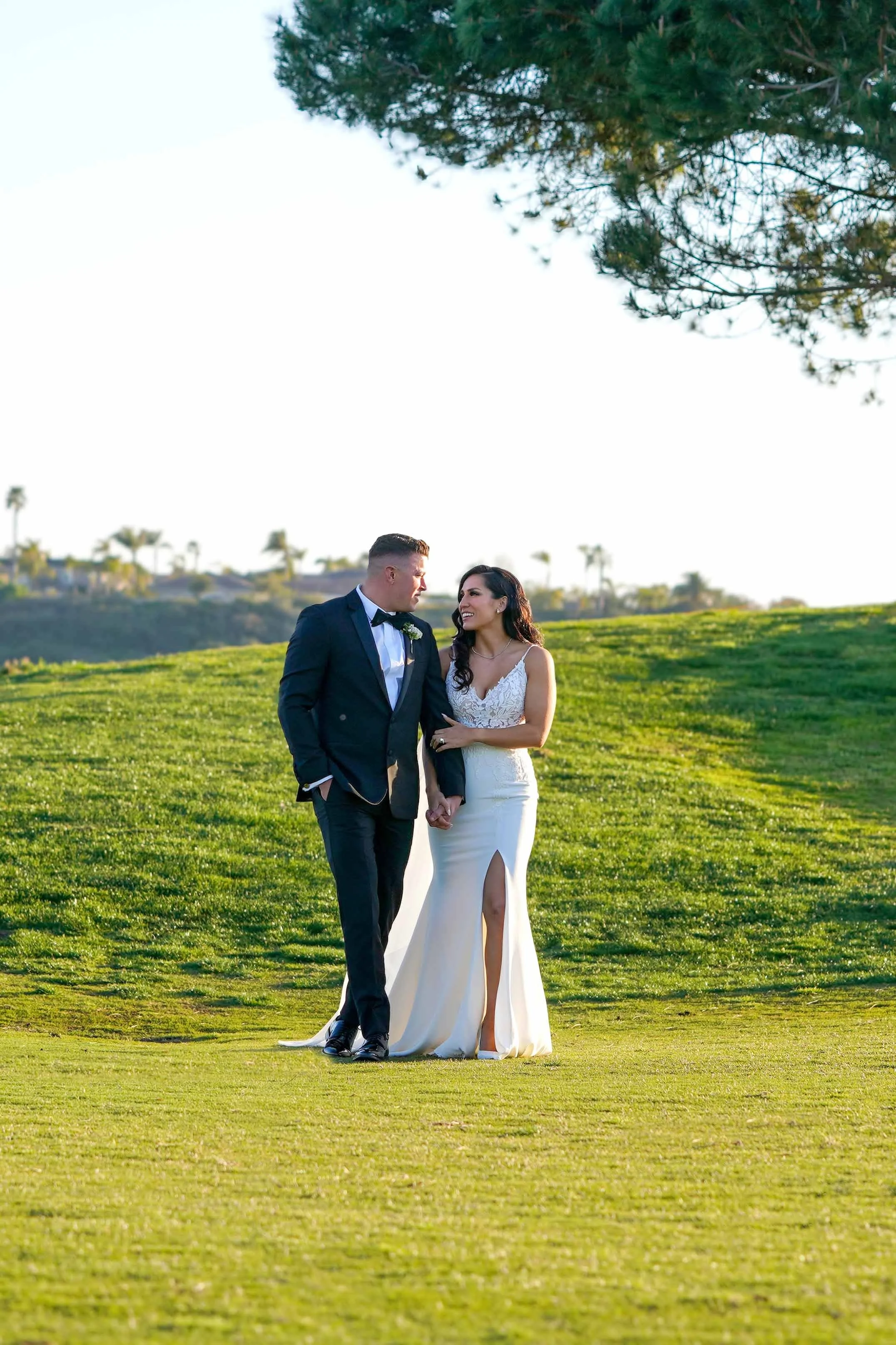 A full-length outdoor photograph of a newly married couple walking hand-in-hand on a grassy hill. The groom wears a dark suit and the bride wears a white wedding dress with a high leg slit, looking at each other. Lush green grass and trees, including