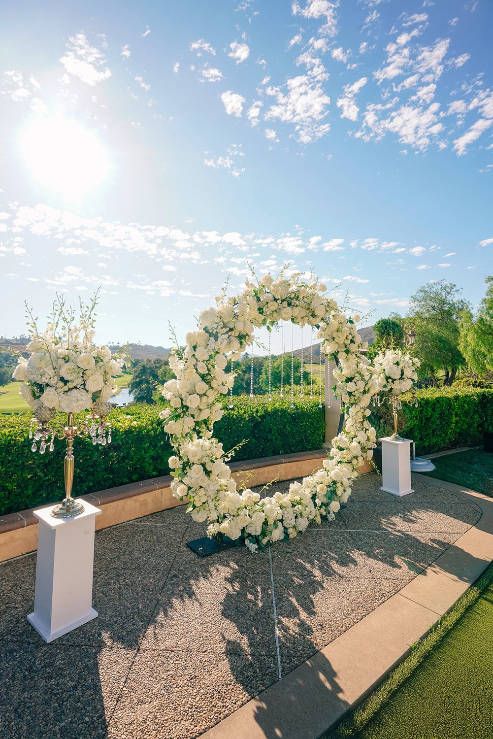 A sunny outdoor photograph of a beautifully decorated wedding ceremony space. The focal point is a large, freestanding circular arch completely covered in white flowers. The arch is flanked by two smaller white floral arrangements on gold stands plac