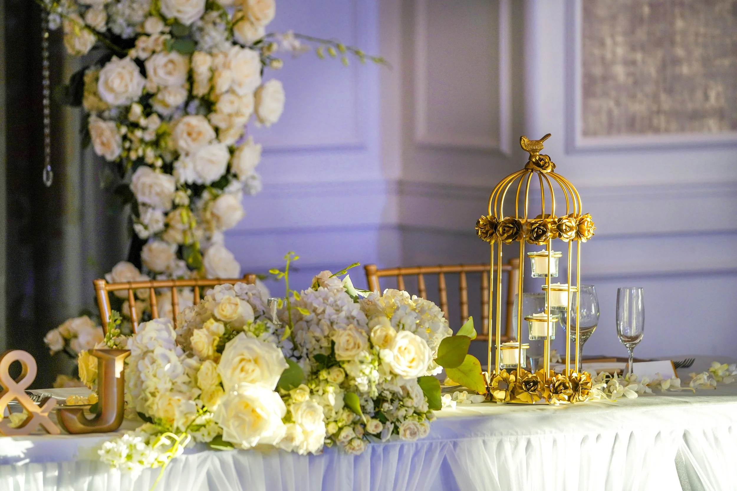 An elegant head table set for an event with a white tablecloth, white floral arrangements, gold "J & J" initials, a gold birdcage centerpiece with candles, and gold chiavari chairs in the background.