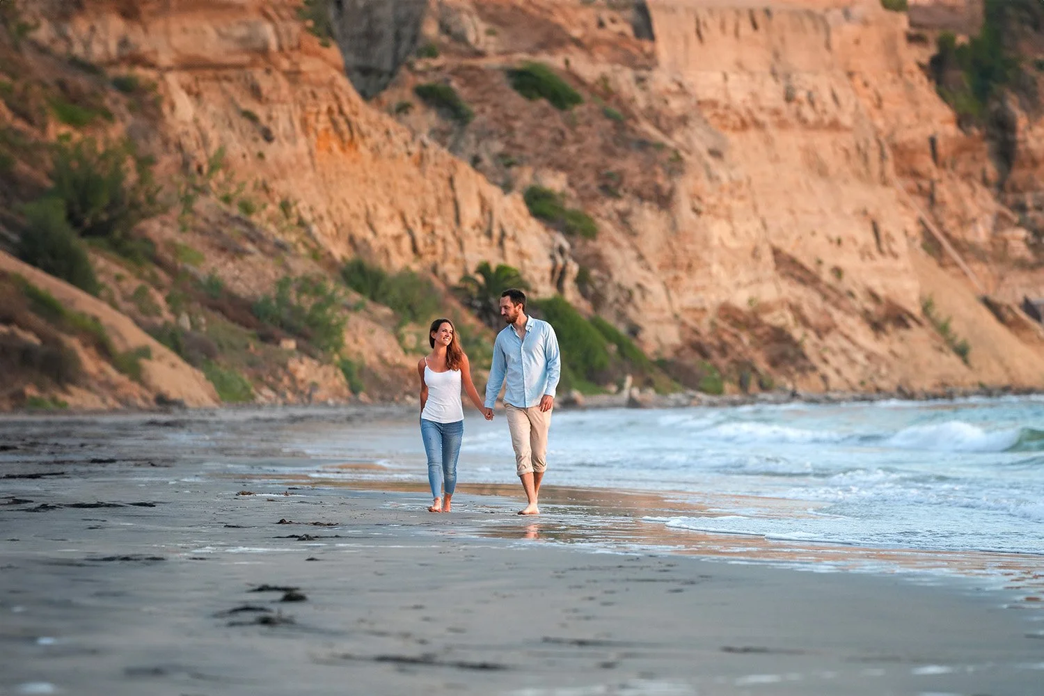 A couple walks hand-in-hand along the beach at sunset, with rocky cliffs and greenery in the background.