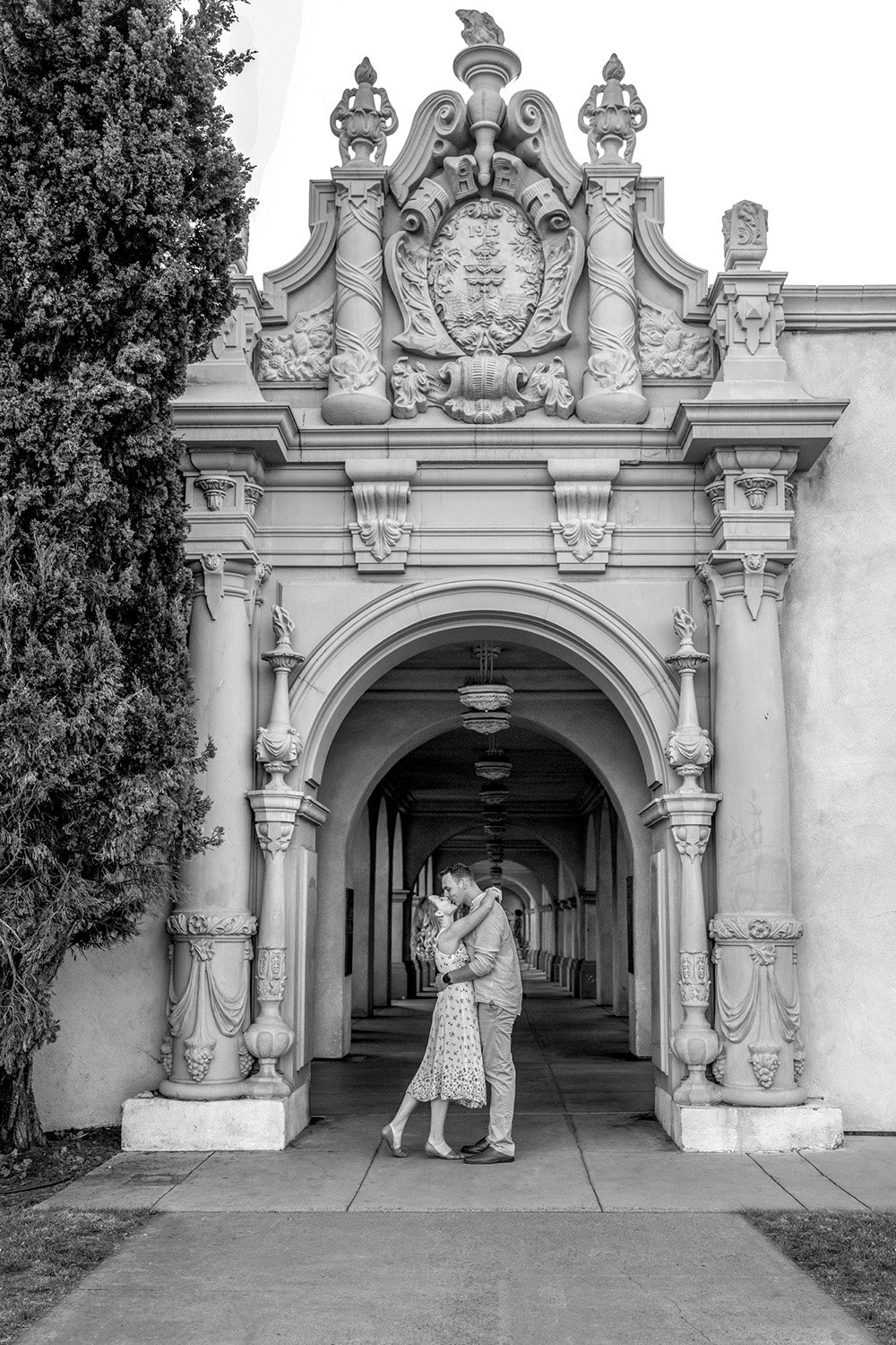 A black and white photograph of a couple embracing and kissing in an arched hallway with Spanish Colonial Revival architecture in Balboa Park. The hallway features ornate stone pillars and arches. A dark tree trunk is visible to the left, and a paved