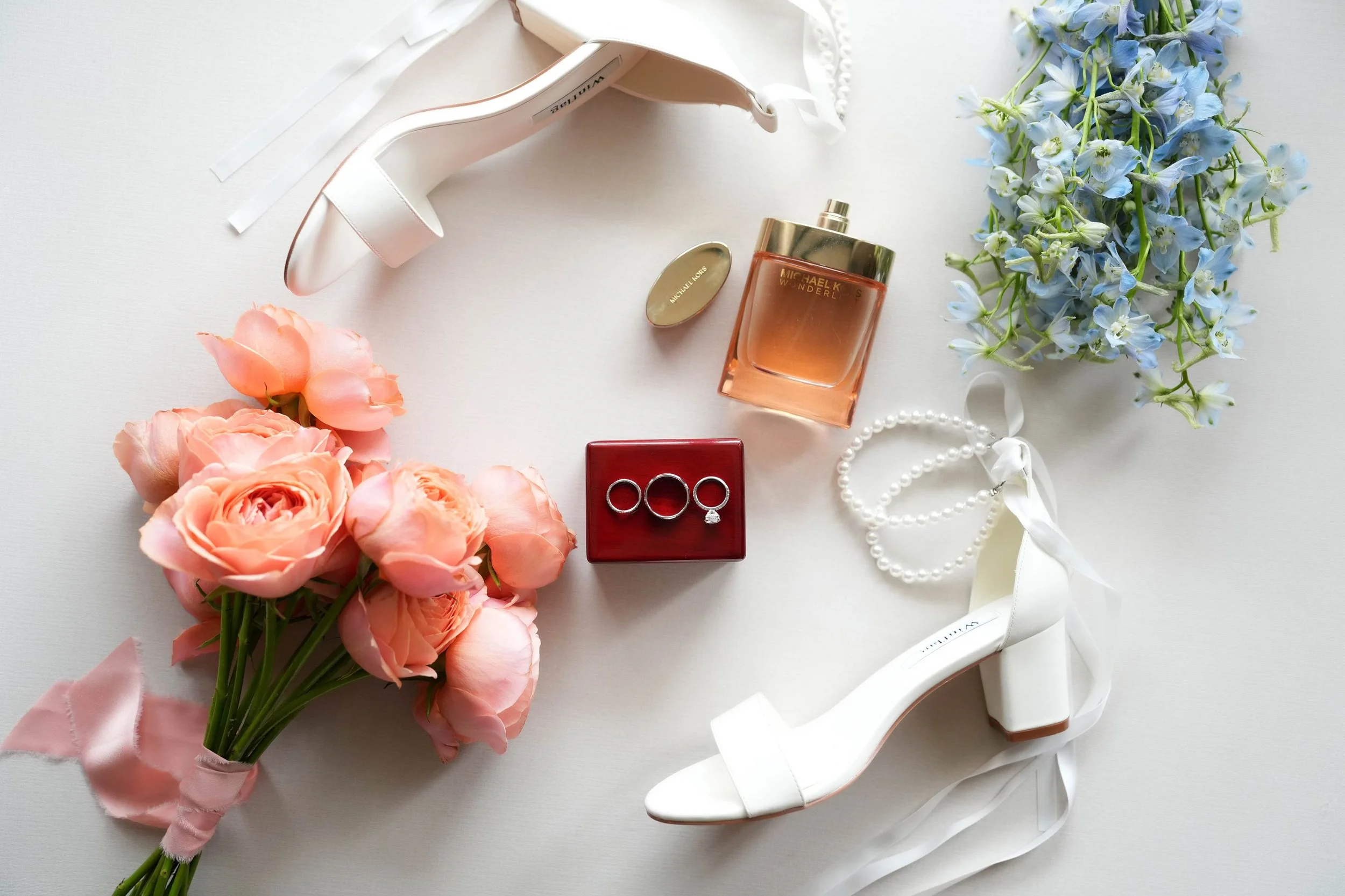 A flat lay photograph of various wedding accessories including white high-heeled sandals, a red box with three wedding rings, a perfume bottle, a pearl necklace, a bouquet of peach roses, and a bouquet of blue flowers.



