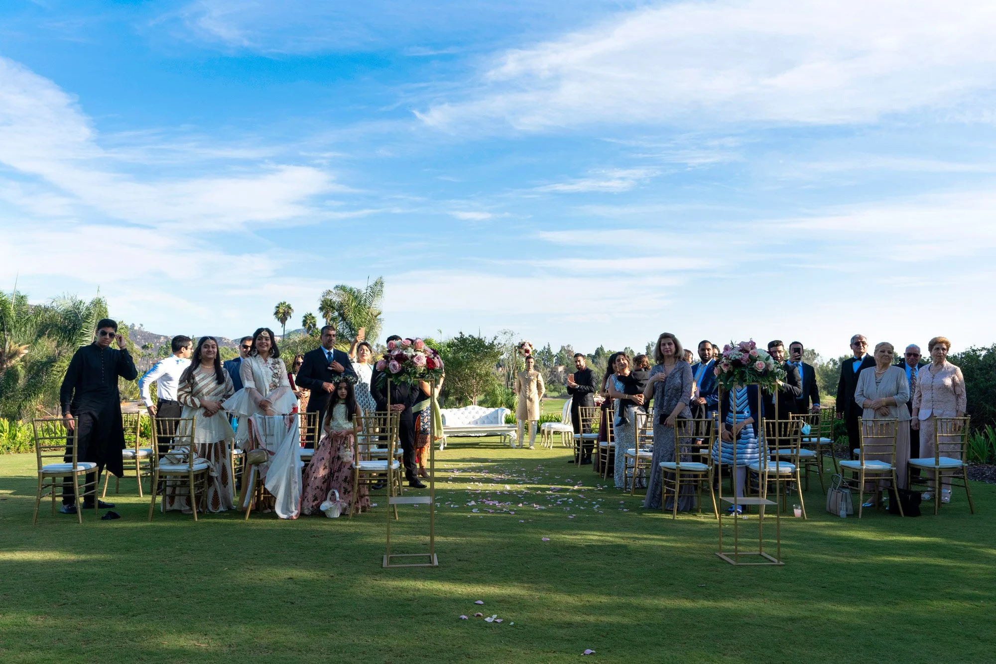 A wide, outdoor photograph of guests attending a bright, daytime wedding ceremony on a grassy lawn. Guests are dressed in a mix of Western and traditional Indian attire and are seated on gold Chiavari chairs. The scene is set under a blue sky with wh