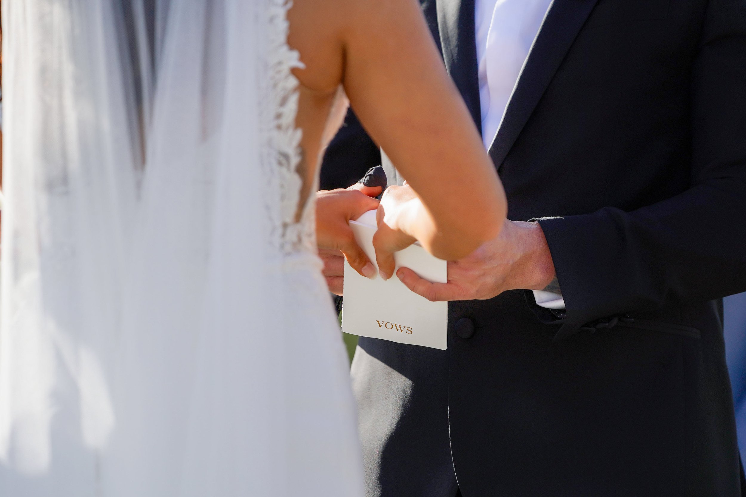 A close-up photograph capturing the hands of a bride in a white dress and a groom in a black tuxedo as they hold a small white booklet labeled "VOWS" during their outdoor wedding ceremony.


