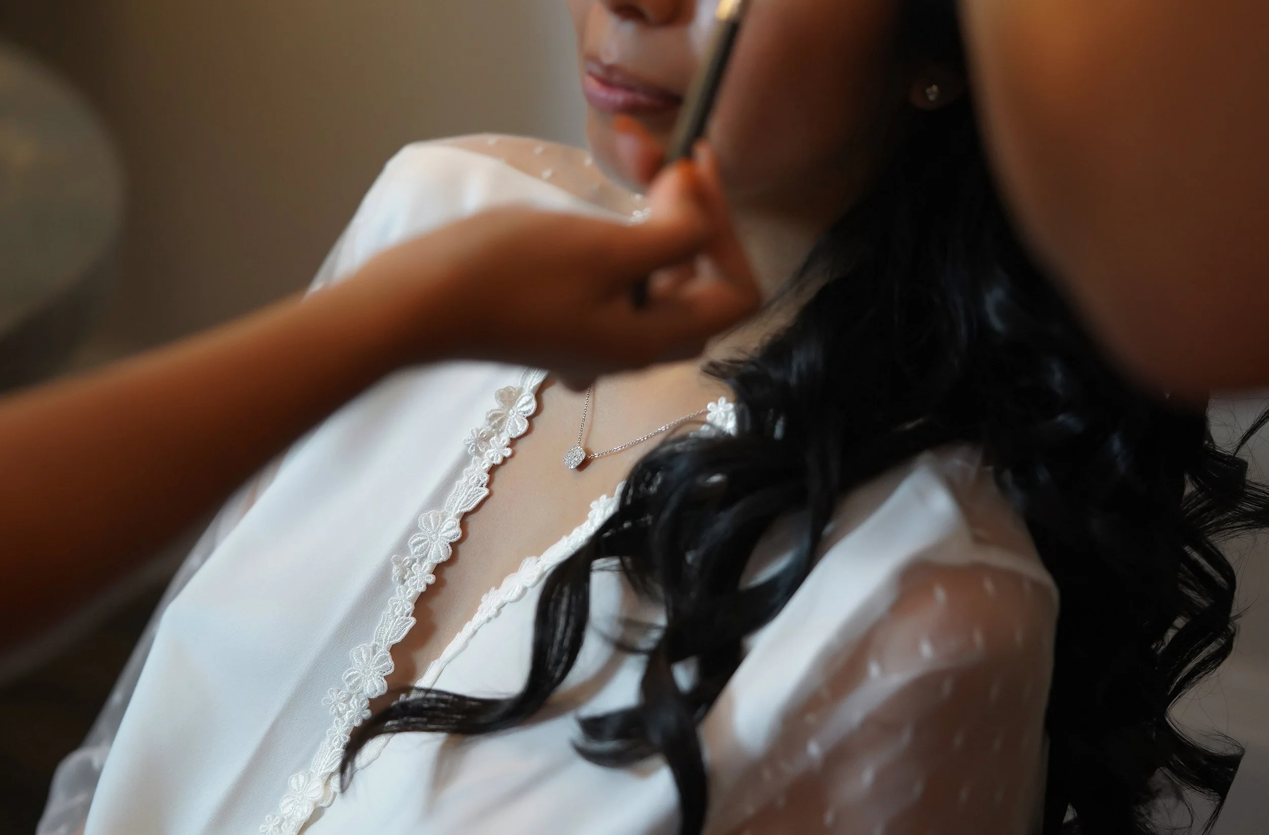 Detailed shot of a bride's wedding day preparation, featuring a sheer swiss dot bridal robe, a diamond necklace, and long dark wavy hair.