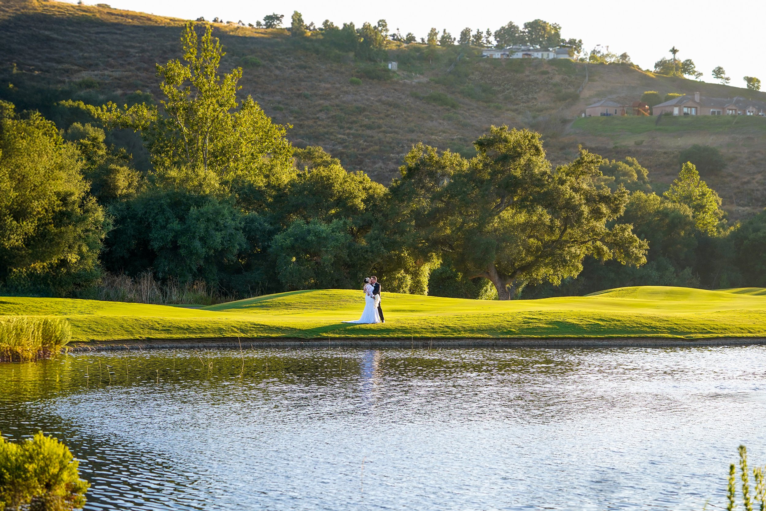 The couple, a bride in a white gown and a groom in a dark suit, are positioned in the middle distance on a manicured grassy hill, in maderas golf club in poway california, which slopes down to a calm pond or small lake in the foreground. Their figure