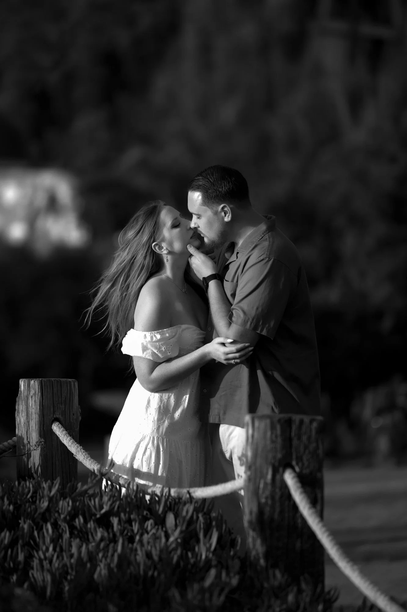 A black and white outdoor photograph of a couple embracing and about to kiss. The woman wears an off-the-shoulder white dress, and the man wears a dark shirt and light-colored pants. They stand near a rope and wooden post barrier in the foreground, a