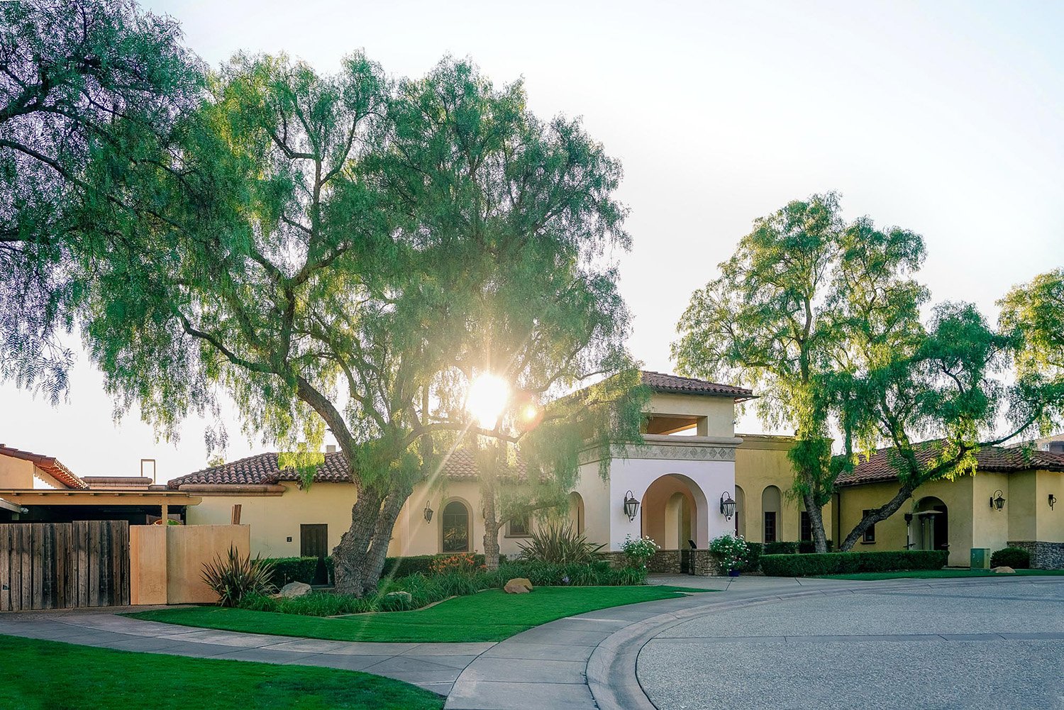 Spanish-style clubhouse building at the Maderas Golf Club, viewed from the end of a paved driveway. A large tree is prominently featured in the foreground and center, with the bright sun shining through its leaves. The stucco building has an arched e