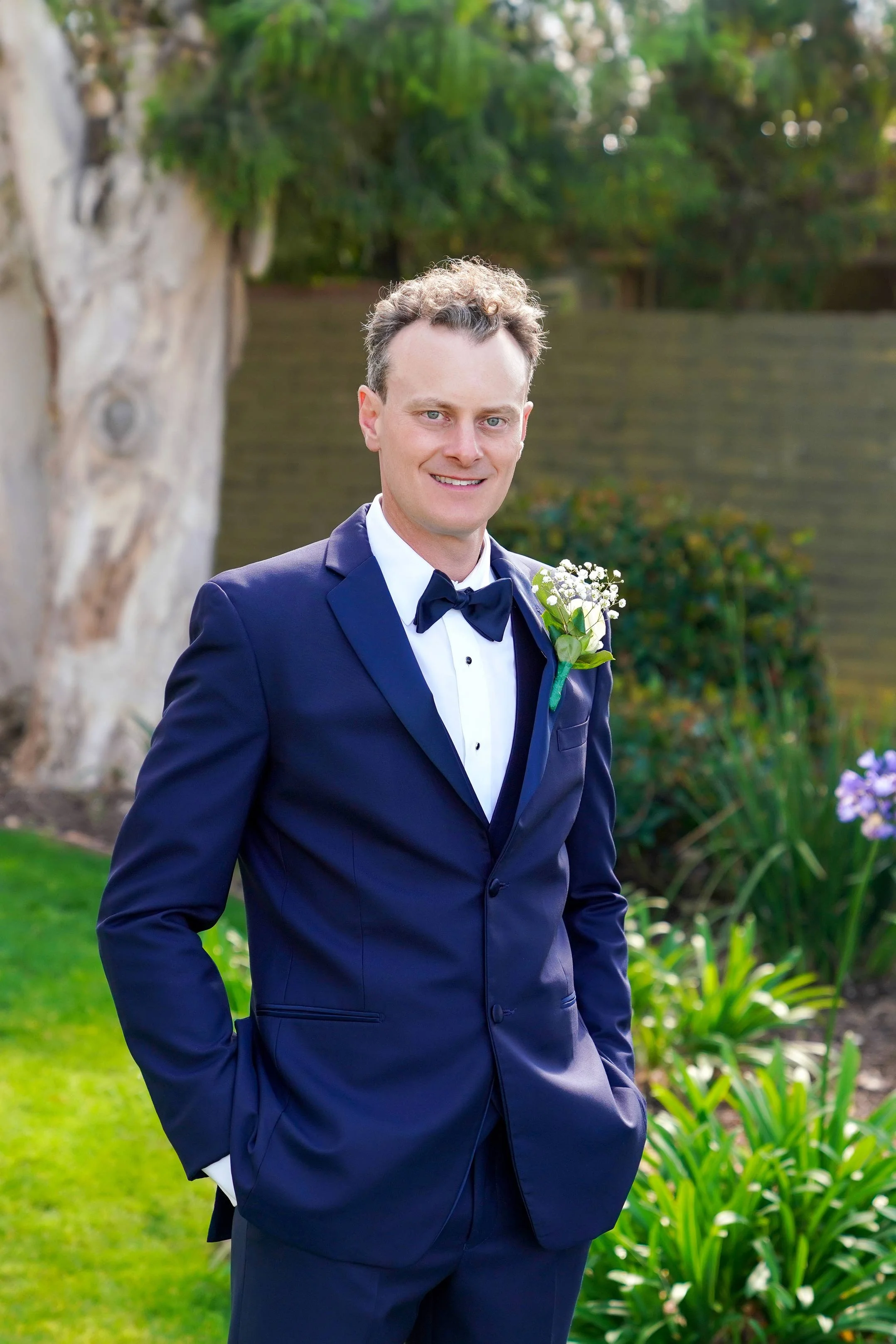 A smiling groom in a navy blue tuxedo with a black bow tie and a white floral boutonniere poses outdoors in a sunny garden setting on Coronado Island.


