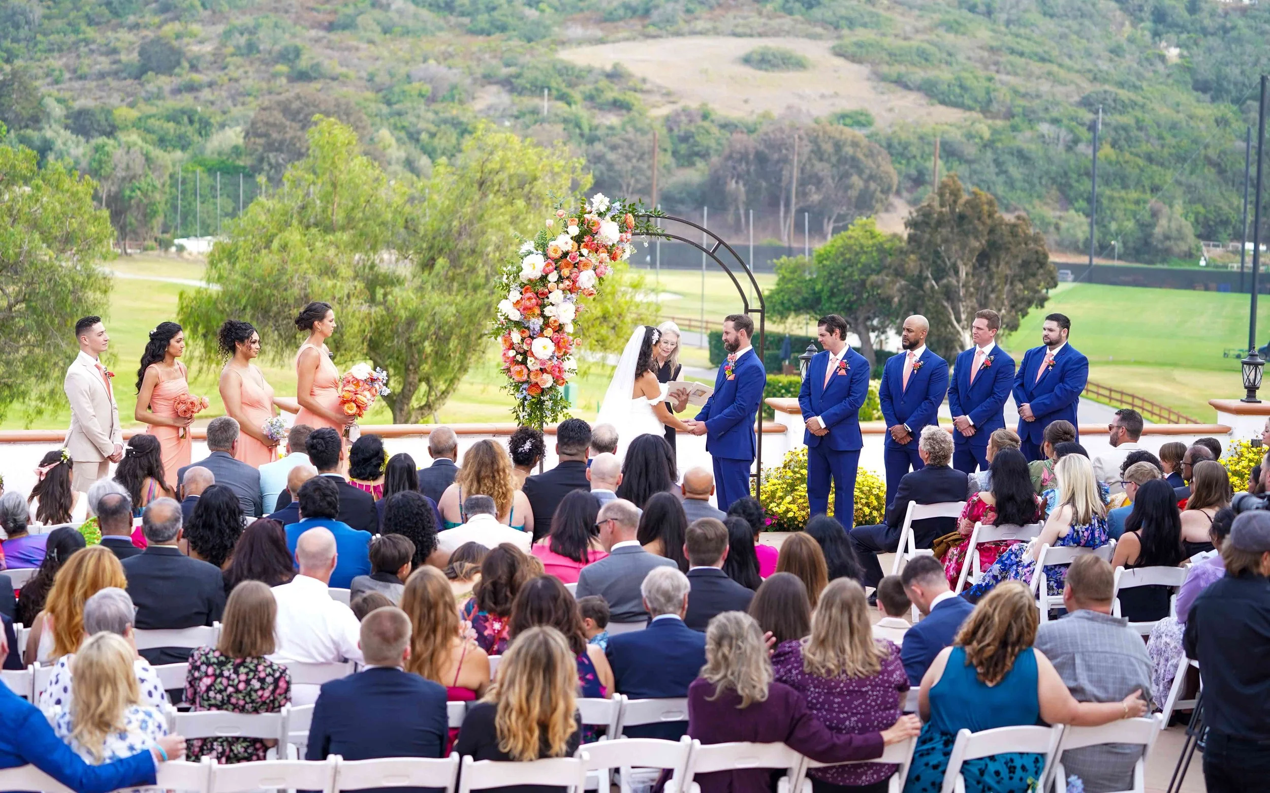 An outdoor wedding ceremony takes place on a green lawn overlooking a hilly landscape, featuring a bride in a white dress and a groom in a navy suit standing at an altar under a floral arch, surrounded by their wedding party and seated guests at Omni