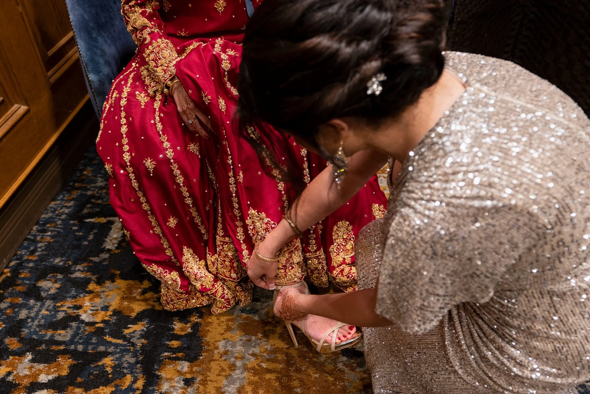 A close-up photograph capturing an indoor moment where a person in an ornate, deep red and gold outfit is seated, and another person in a shimmering gold dress is kneeling to help them with a gold strappy high heel shoe. The scene takes place in a ro