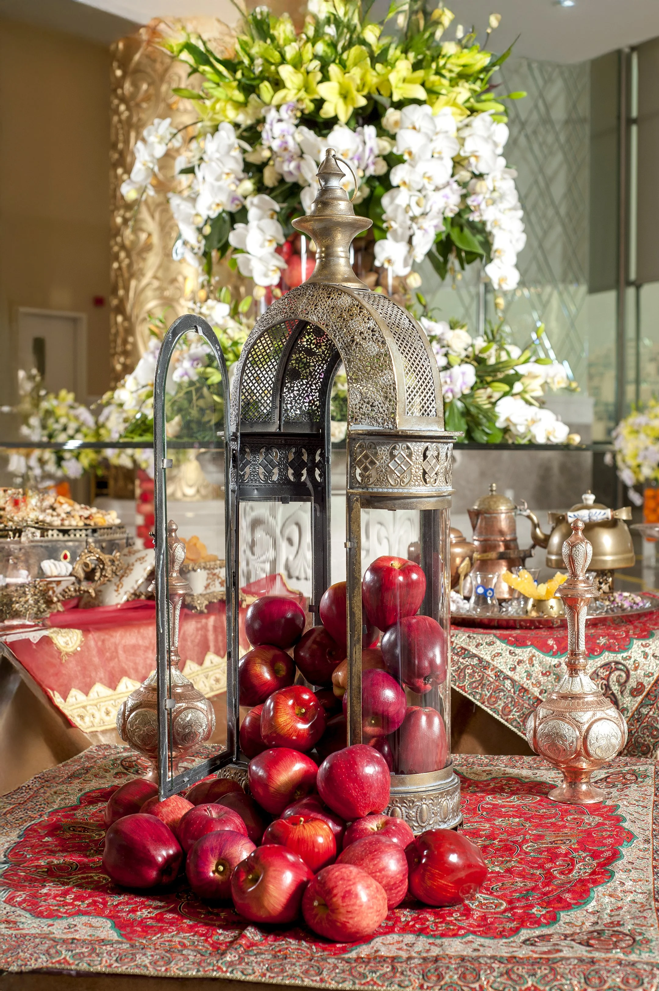 An ornate metal lantern filled with red apples and surrounded by more apples, displayed on a traditional red patterned tablecloth as part of a Yalda Night or Iranian celebration table setting, with silver teapots and white floral arrangements in the 