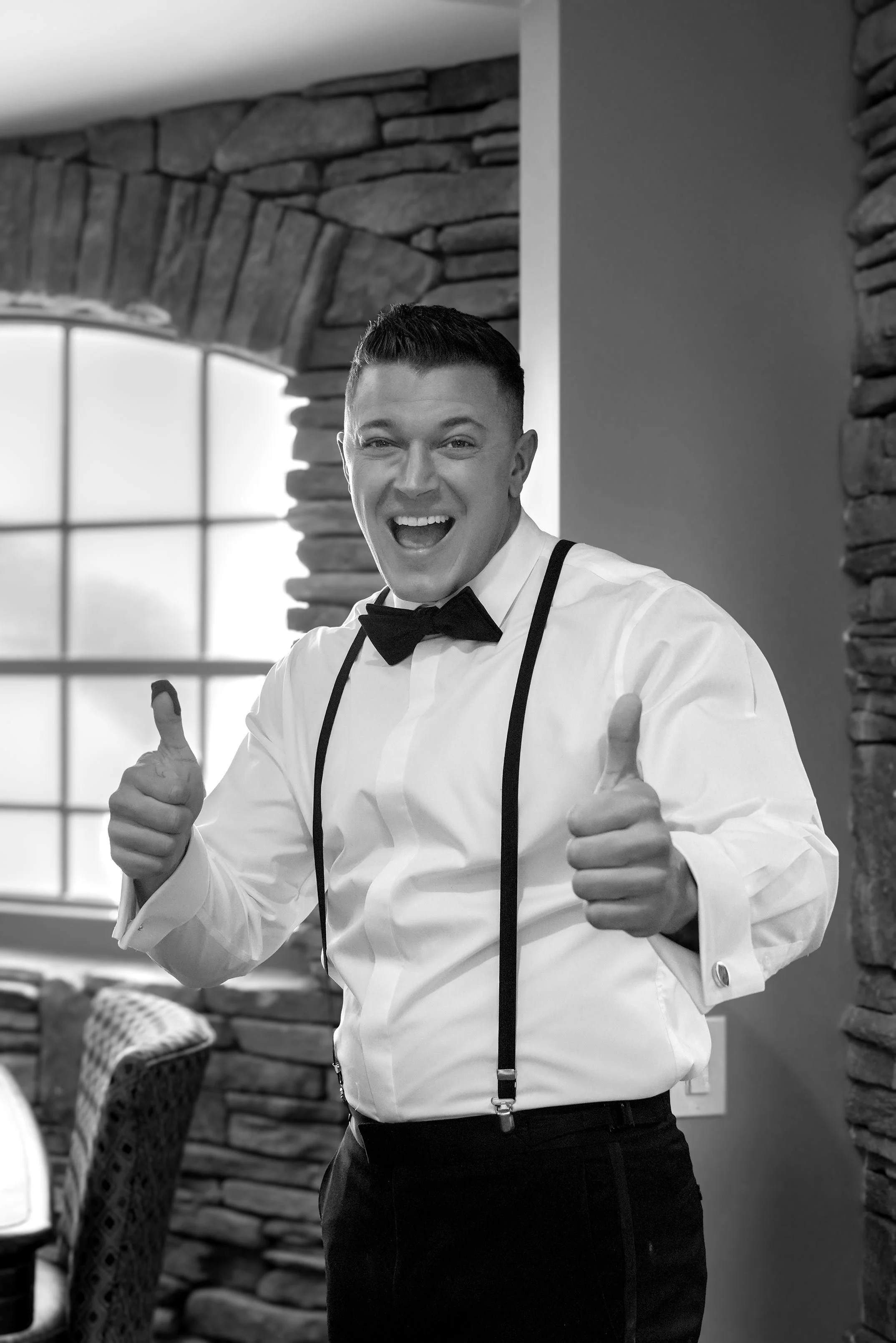 A black and white photograph of a groom in a white dress shirt, black suspenders, and a black bow tie smiling widely at the camera and giving two thumbs up. The man stands indoors in a room featuring stone walls and a window with a grid pattern in th