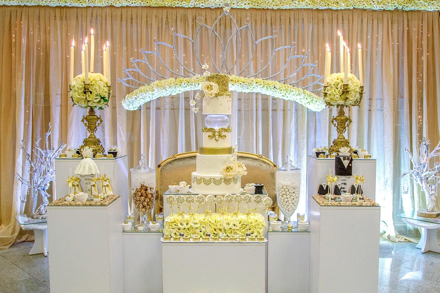 An elaborate, formal wedding dessert table featuring a multi-tiered white cake with gold accents, flanked by white floral arrangements on gold stands with tall candles, assorted treats in clear jars, and decorative white display items.