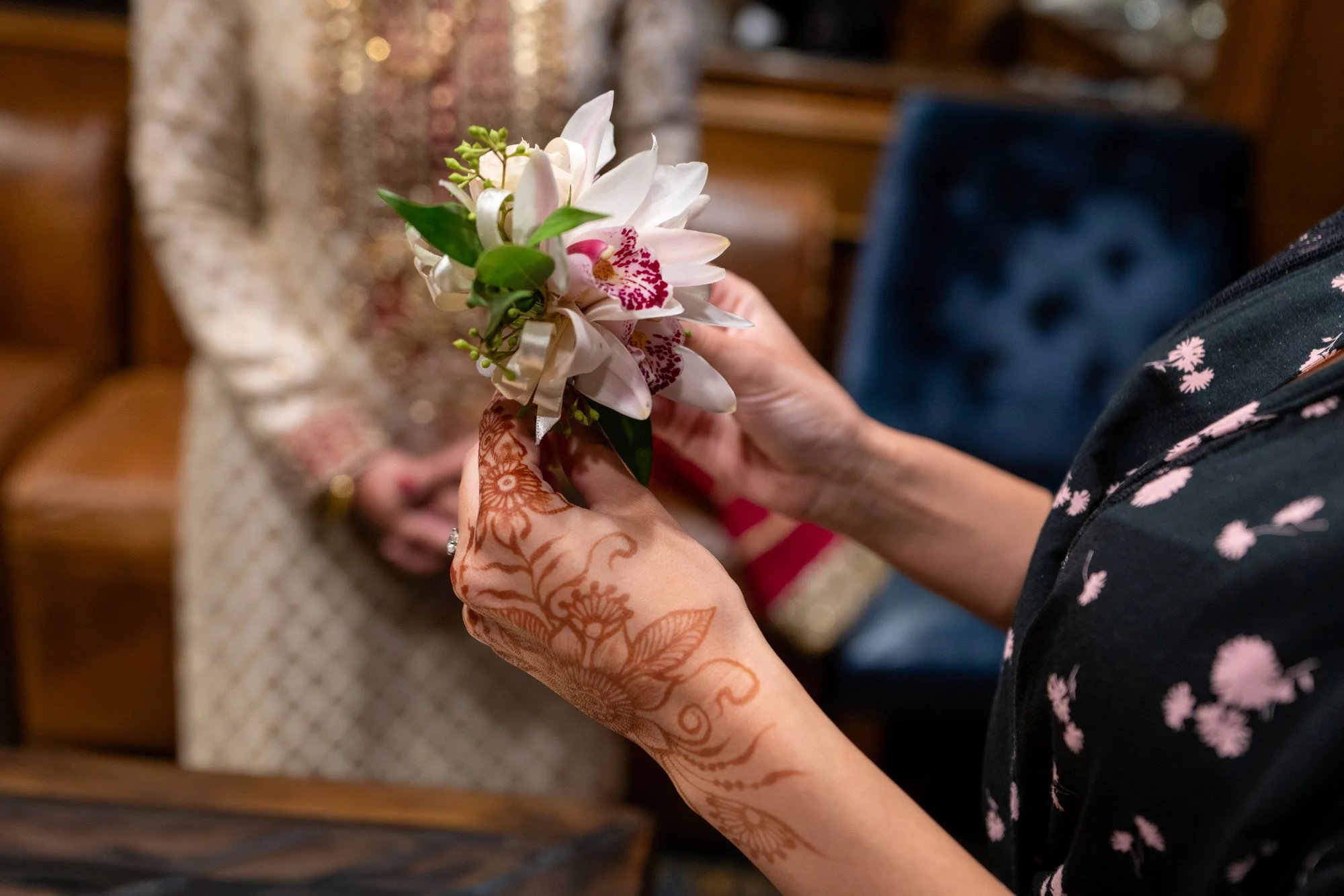 A close-up photograph of hands with elaborate henna tattoos holding a small boutonnière or corsage made of white orchids with pink centers. A person in traditional Indian formal wear (sherwani) stands in the blurred background.


