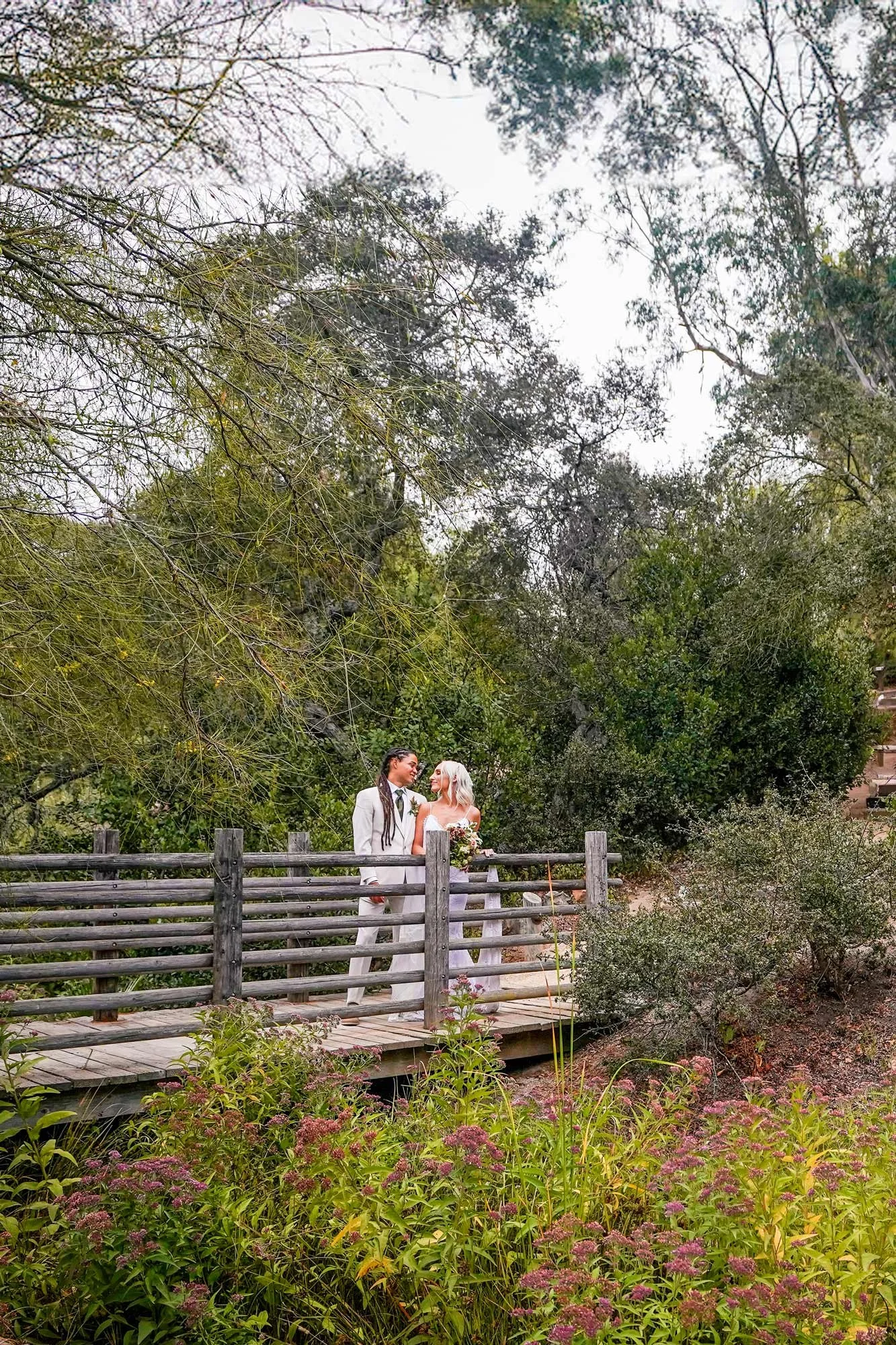 A photo of a newly married couple embracing and looking into each other's eyes on a rustic wooden bridge, surrounded by lush green trees and plants. The location appears to be the Leo Carrillo Ranch Historic Park in Carlsbad, California.


