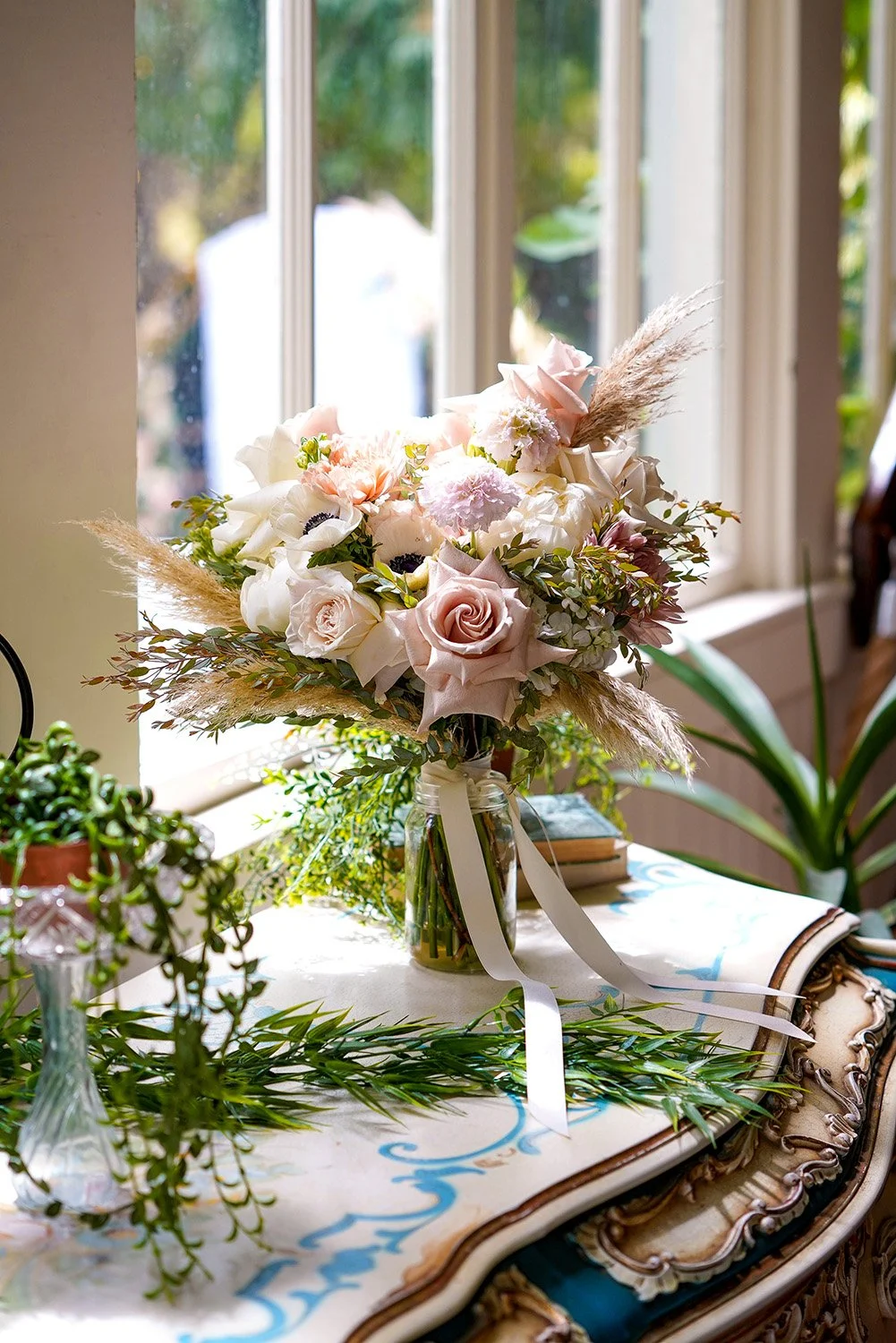 A rustic-style wedding bouquet featuring dusty rose, peach, and white roses with pampas grass, displayed in a clear glass jar on an ornate table near a window.
