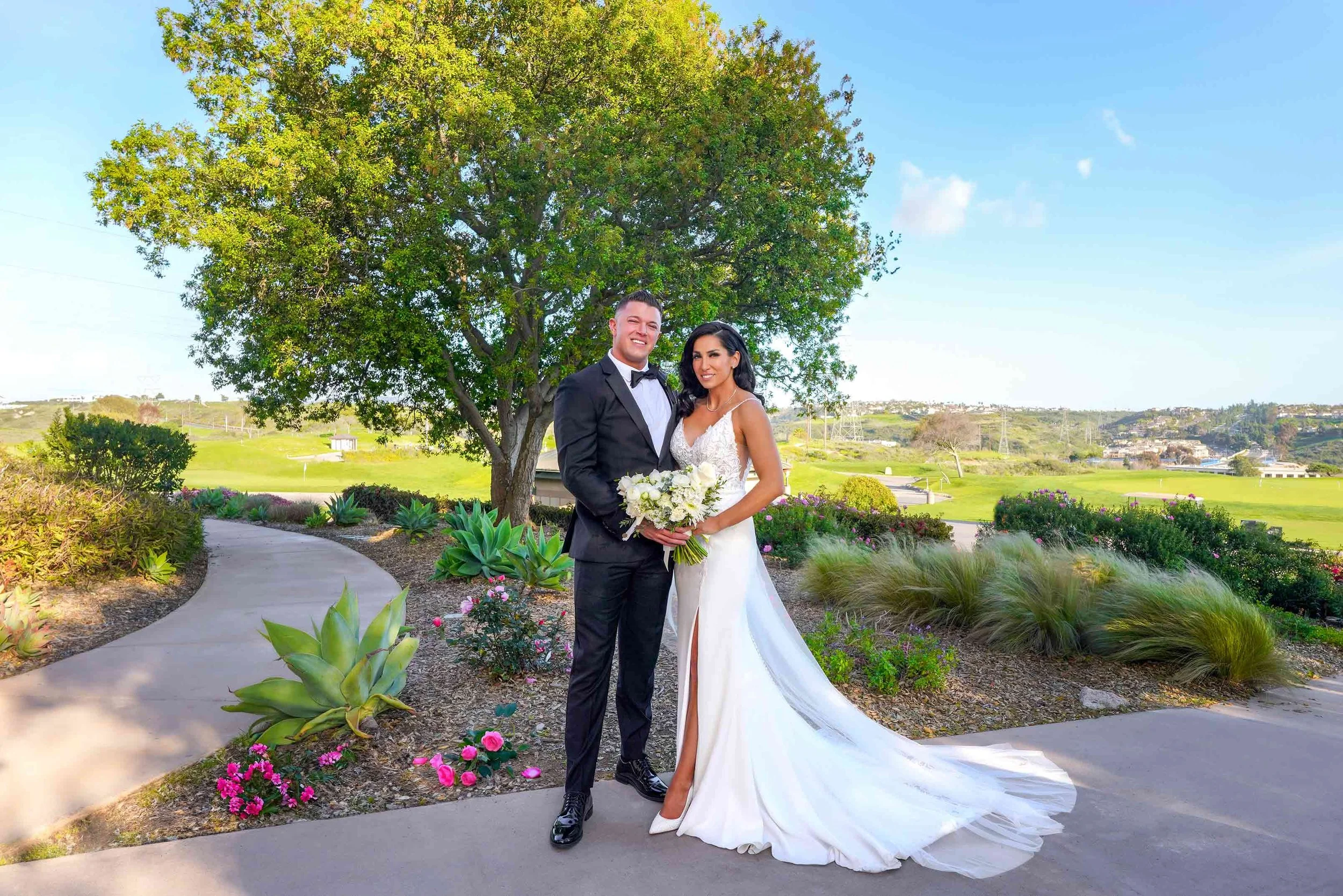 A newly married couple in a tuxedo and white dress poses for a portrait photo on a paved pathway with a large green tree and a golf course landscape in the background on a bright, sunny day in San Diego.



