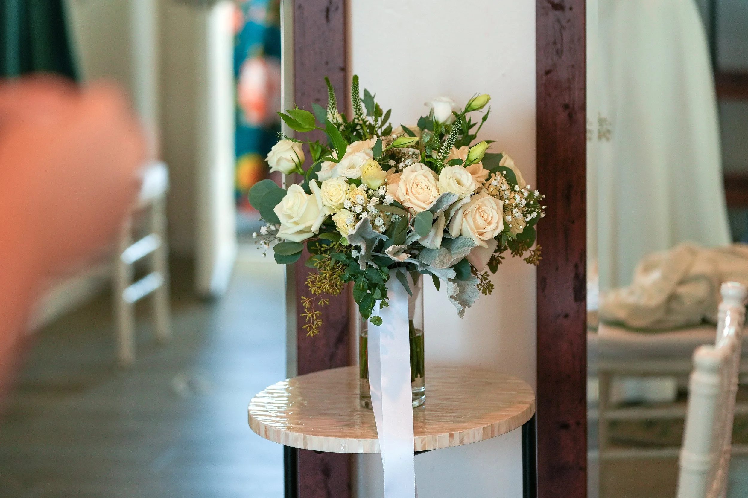 A bridal bouquet featuring white and peach roses, eucalyptus leaves, and baby's breath, tied with a long white ribbon and displayed in a clear glass vase on a round, mother-of-pearl or shell-inlay side table.