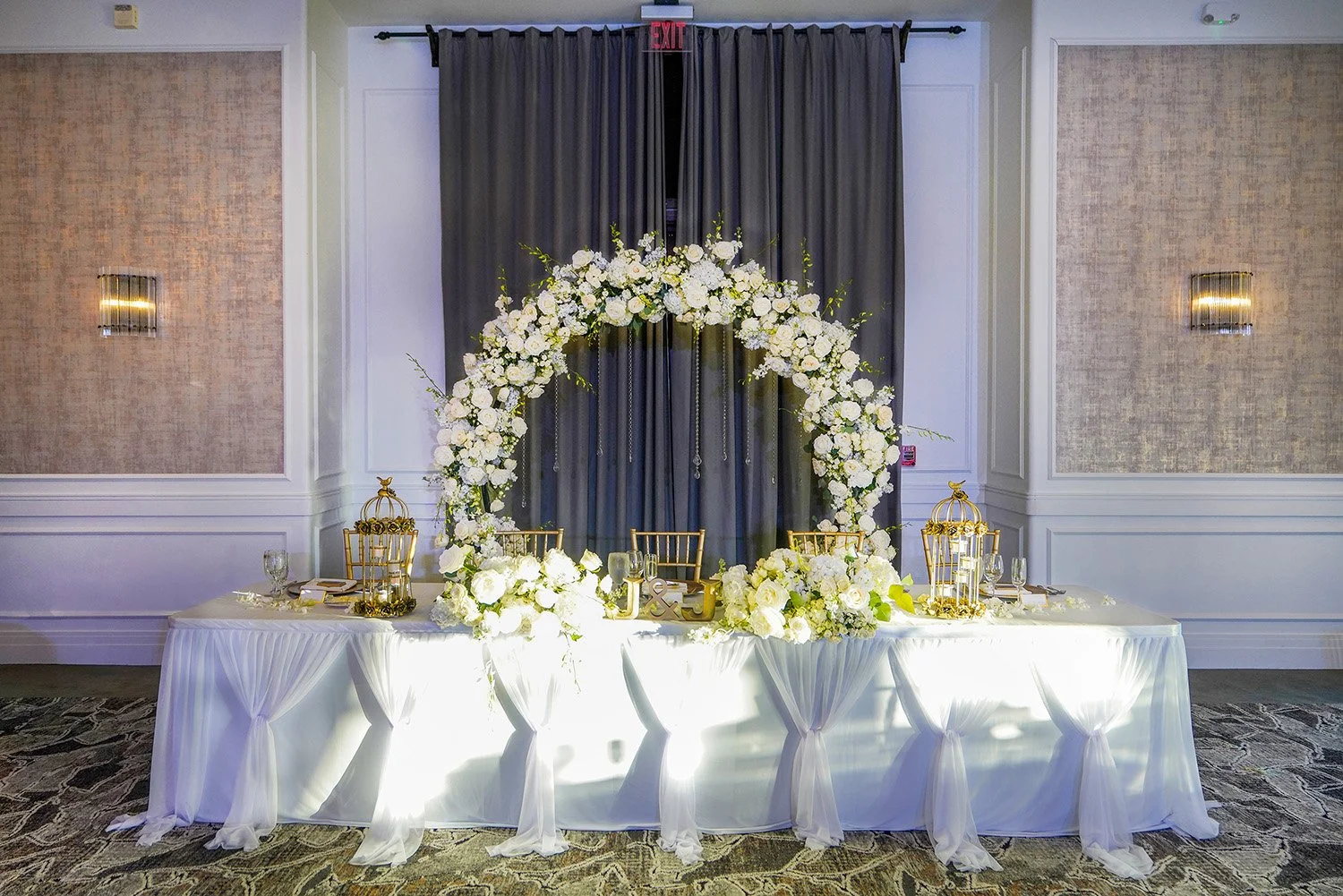 The long table is covered in a white tablecloth with a draped white sheer fabric treatment in the front. The tabletop is adorned with additional arrangements of white flowers and several decorative gold-toned birdcages used as centerpieces. Two gold 
