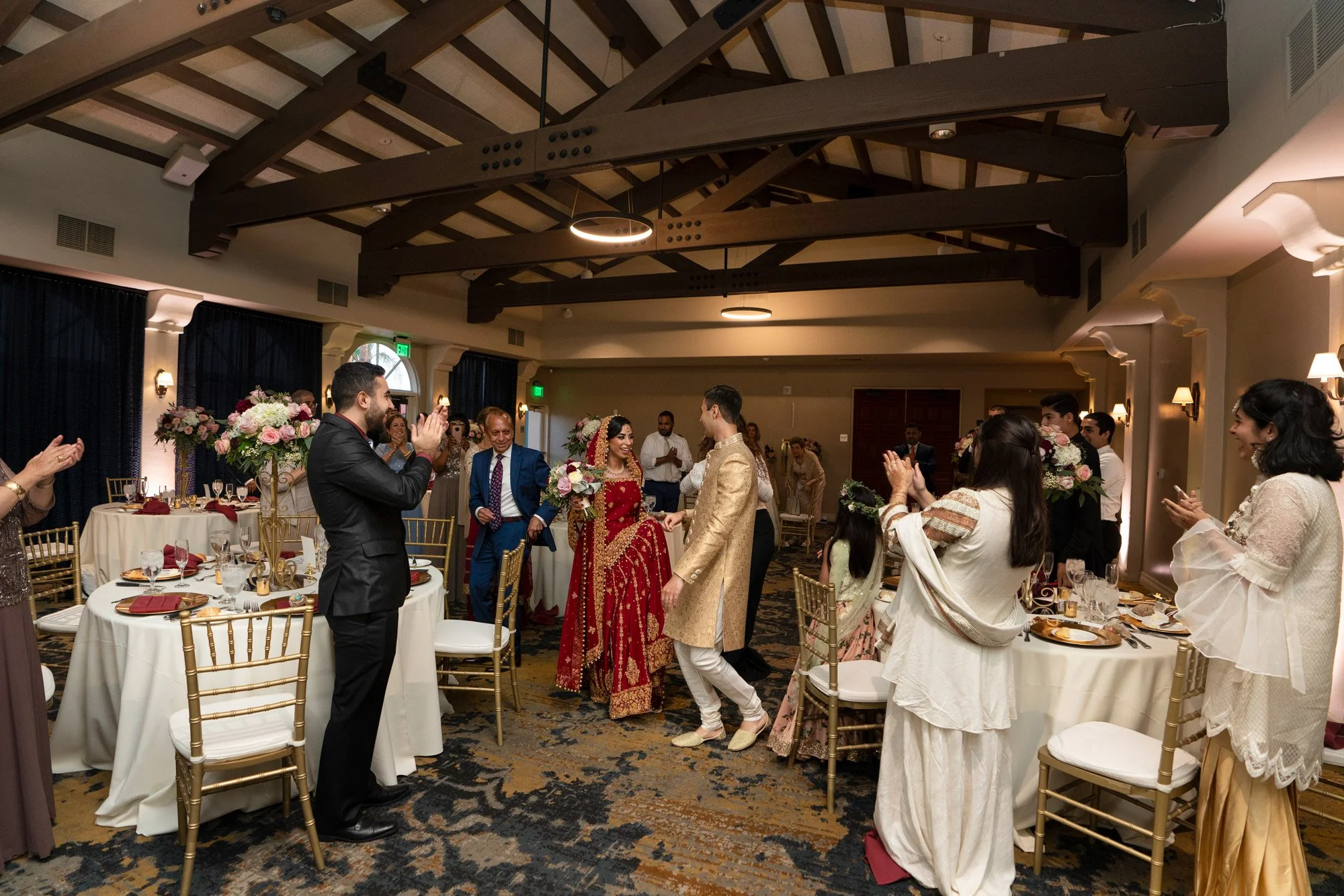 An indoor photograph capturing the wedding reception as the newly married couple is welcomed by guests. The bride is wearing an ornate, deep red lehenga with gold embroidery, and the groom is in a gold sherwani. They are walking down an aisle between
