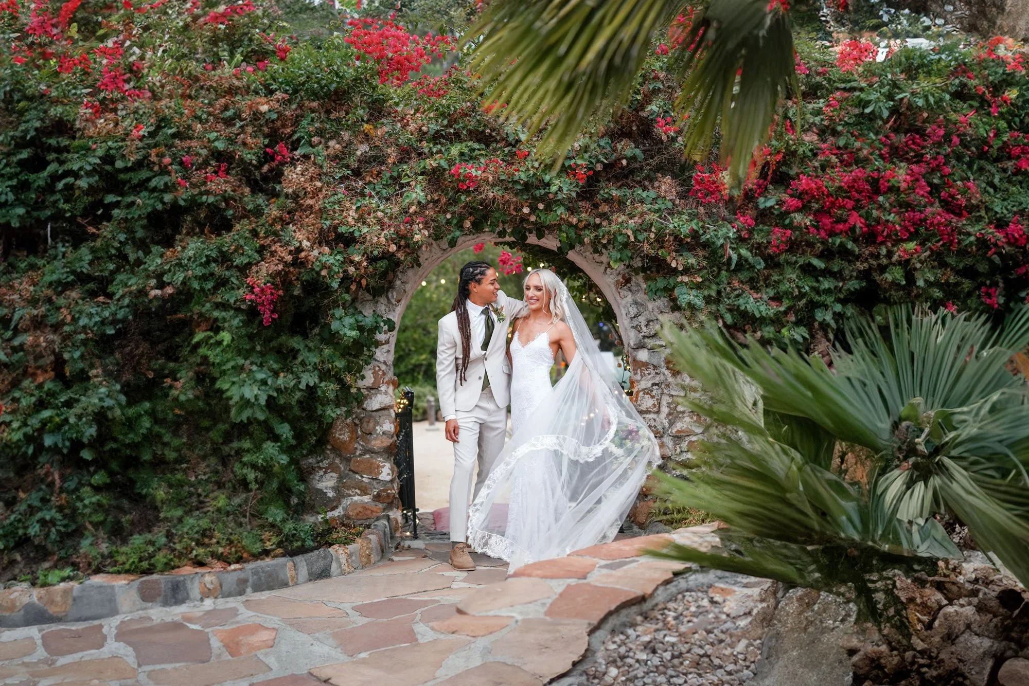A photo of a newly married couple walking together through a stone archway covered in lush green foliage and bright red flowers, with one wearing a cream-colored suit and the other in a white lace dress with a long, flowing veil.


