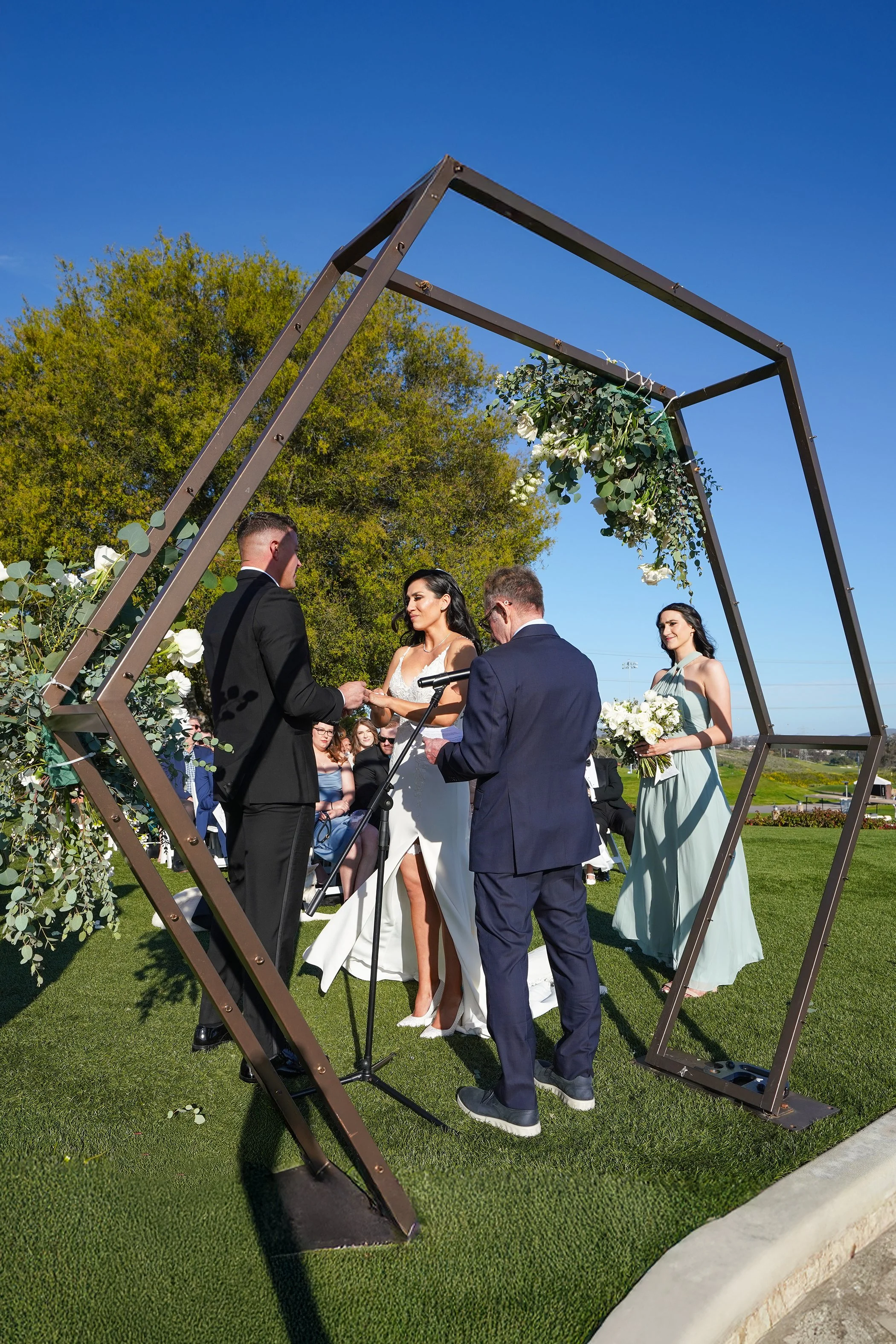 An outdoor wedding ceremony takes place on a bright, sunny day under a clear blue sky. A bride and groom stand facing each other beneath a modern, hexagonal wooden arch decorated with white roses and eucalyptus.
The bride wears a white, floor-length 