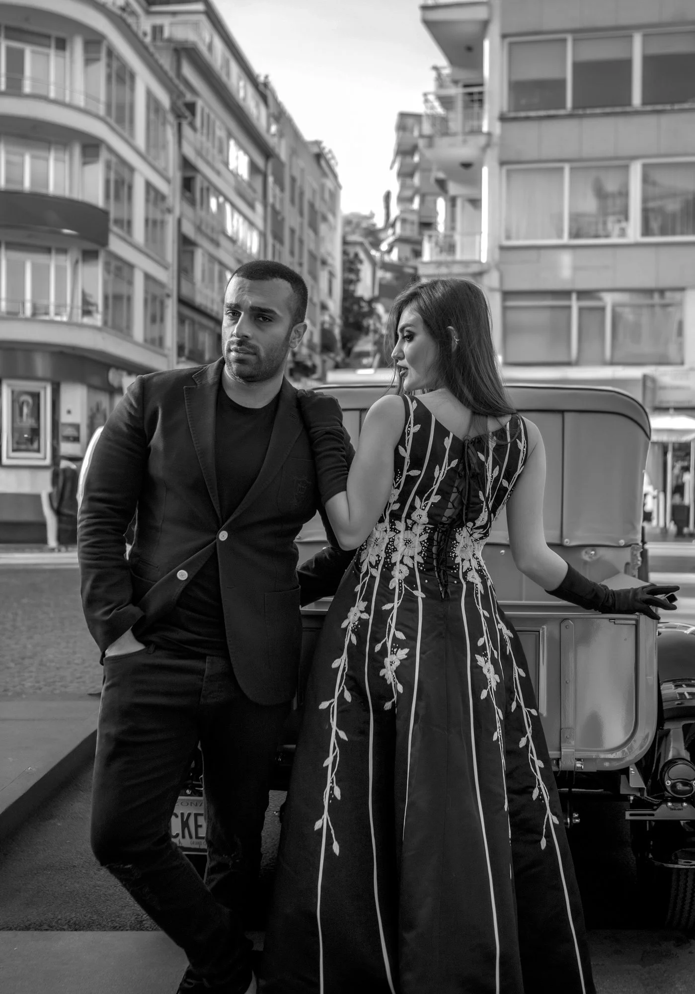 A man and woman in stylish clothing relax on a city street, with multi-story buildings in the background.
