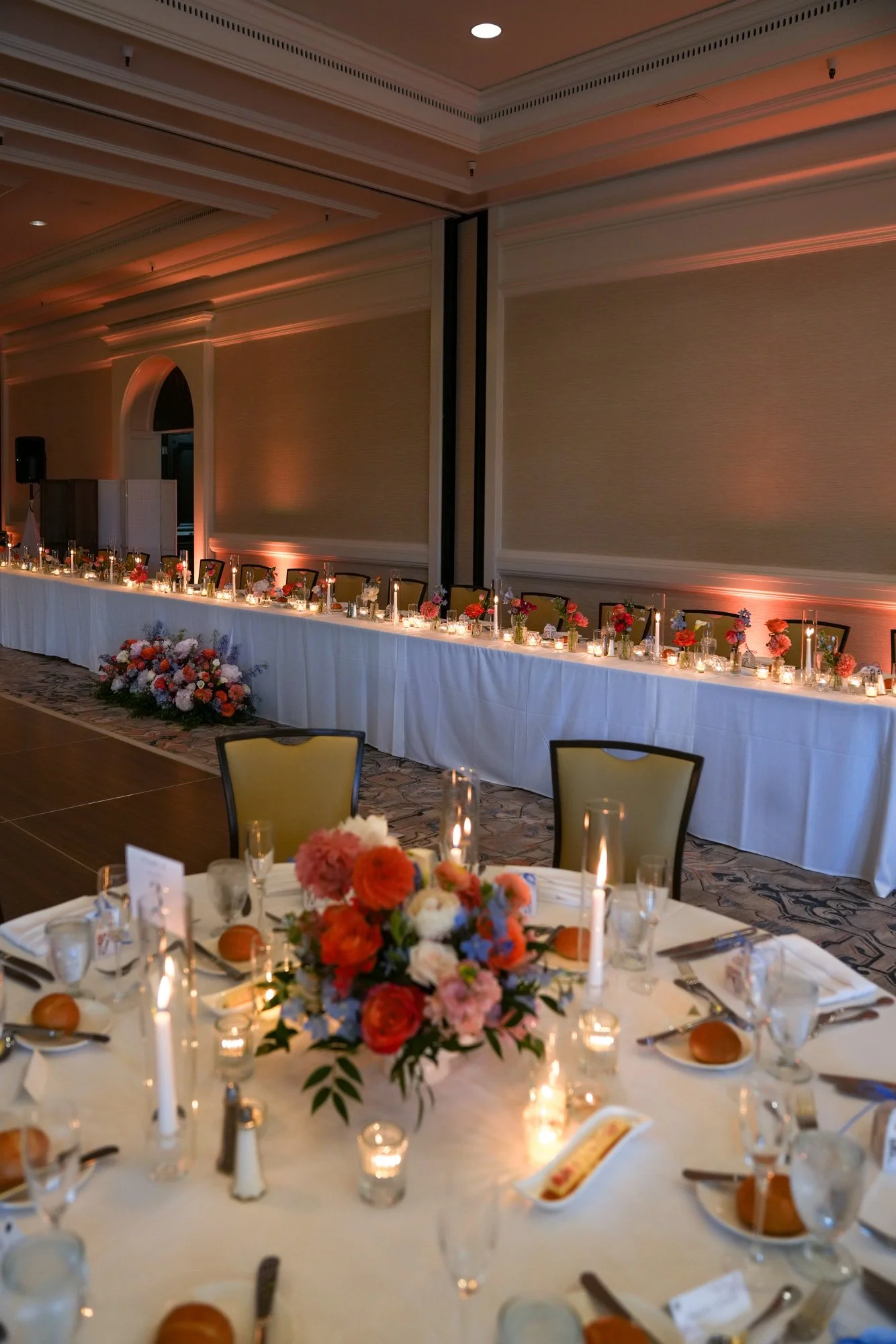 An elegant indoor banquet hall set up for an evening event, featuring round and long tables with white tablecloths, warm candlelight, colorful floral centerpieces with orange, red, and blue flowers, and upholstered chairs.