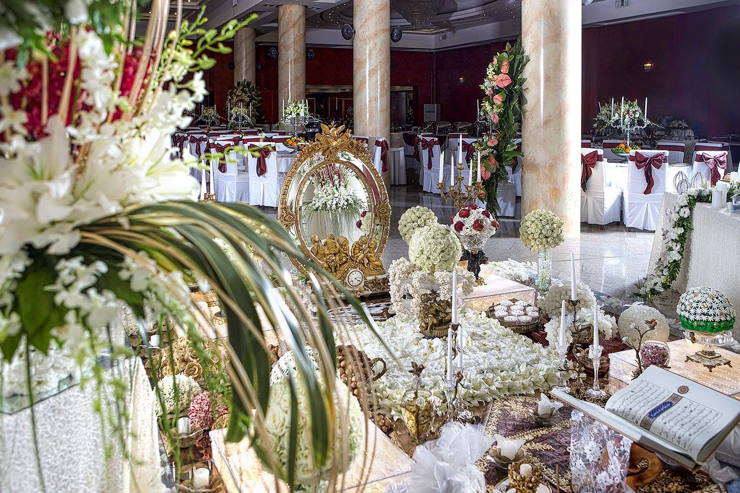 An elaborate, traditional Persian wedding table setting (Sofreh Aghd) featuring an ornate gold mirror, candelabras, a holy book, nuts, sweets, and extensive white floral and gold decorations in a formal banquet hall.