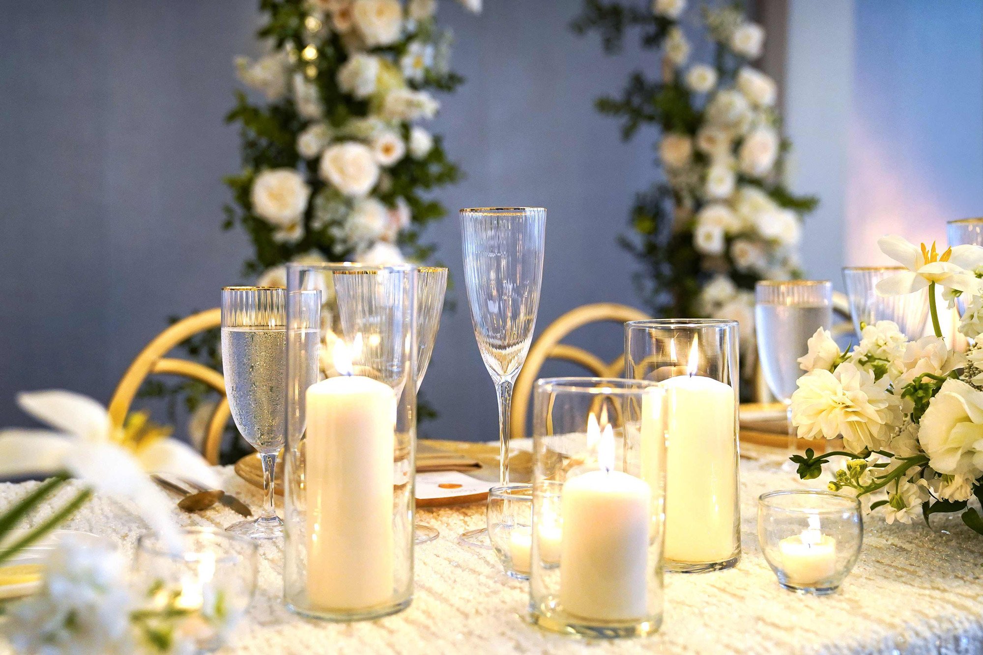 A close-up photograph of an elegantly set dining table for a formal event. The table features a white textured tablecloth, gold-rimmed glasses for water and champagne, and several white pillar candles in glass hurricanes or votives providing a warm g