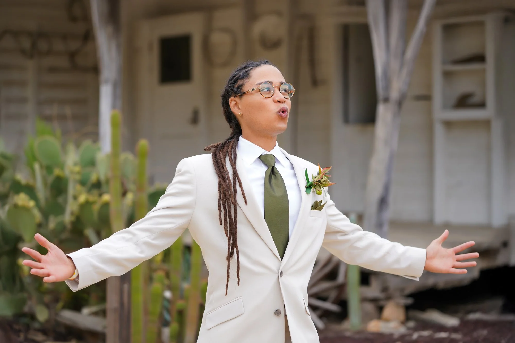 Groom wearing a cream-colored suit, olive green tie, and glasses, standing outdoors with their arms open wide and mouth open as if singing or speaking passionately, in front of an old white building with cacti in the foreground.