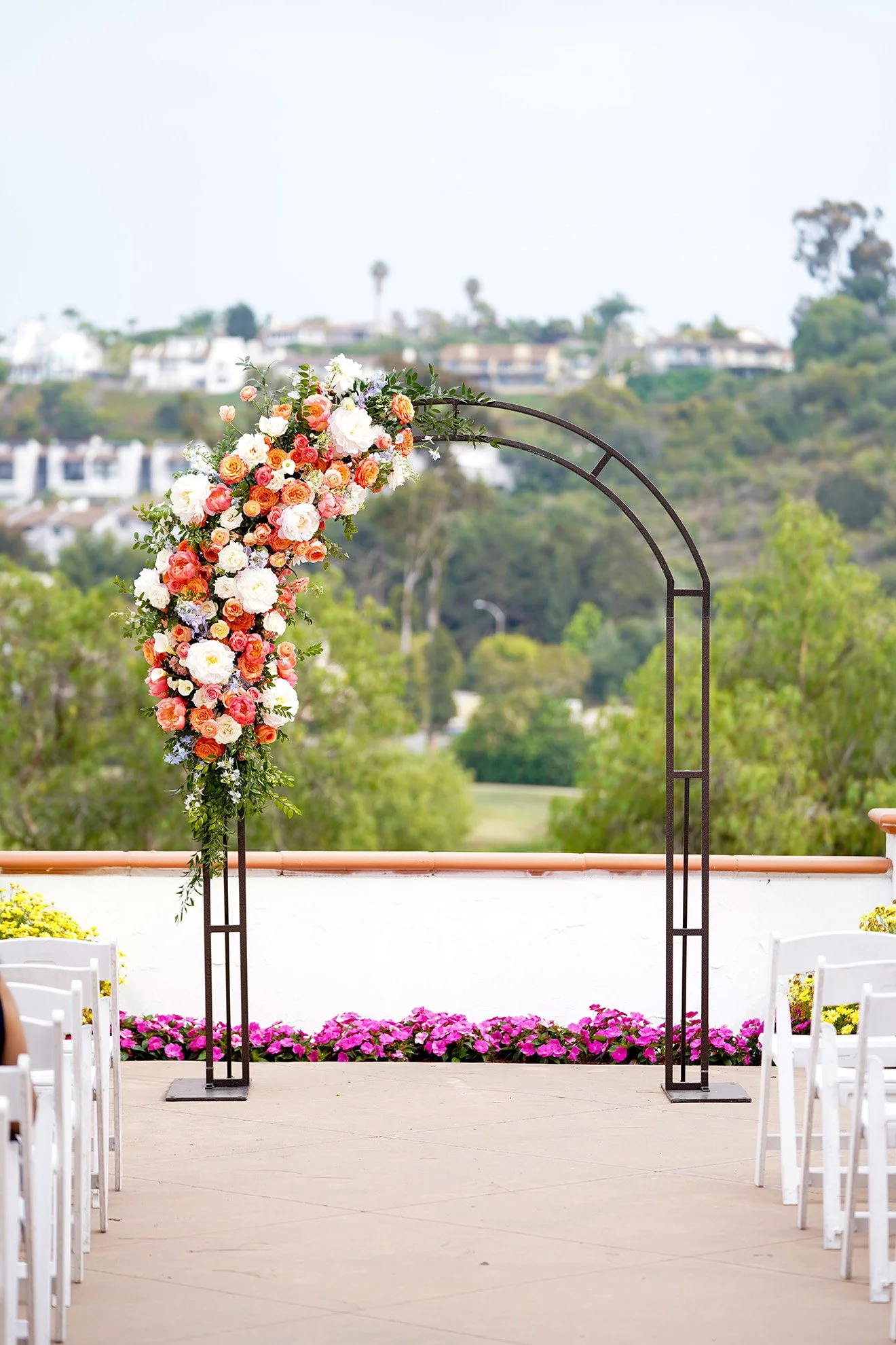 A black metal arch adorned with orange, white, and peach flowers, set up for an outdoor wedding ceremony on a patio overlooking green hills.
