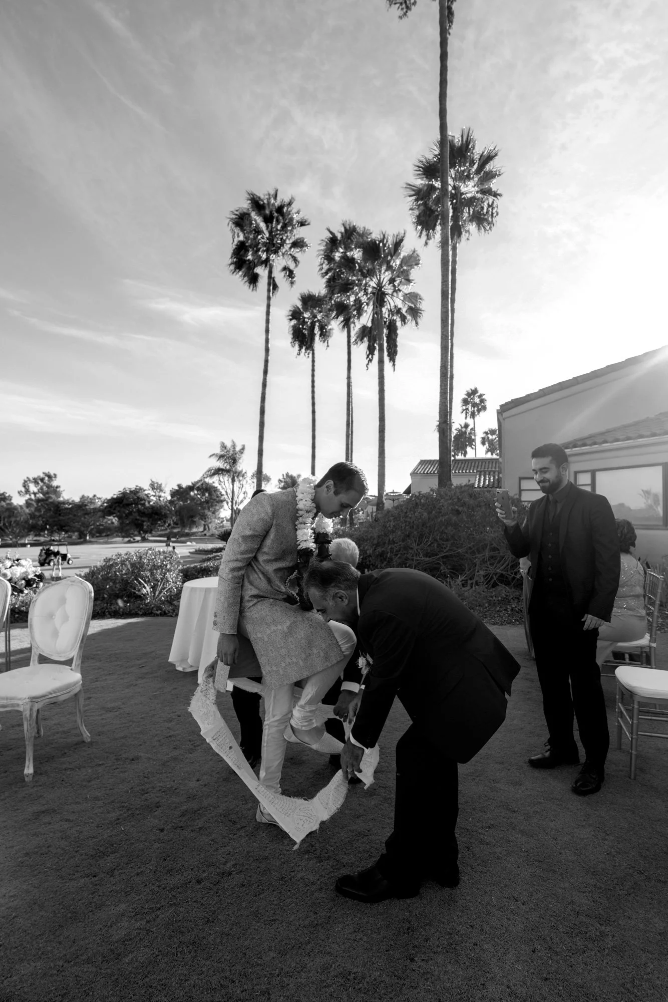 A black and white outdoor photograph capturing a moment during a wedding event. A person in a gold sherwani and garland is seated while another person in a dark suit kneels and appears to be tying a white wrap or ribbon around their leg or shoe. A th