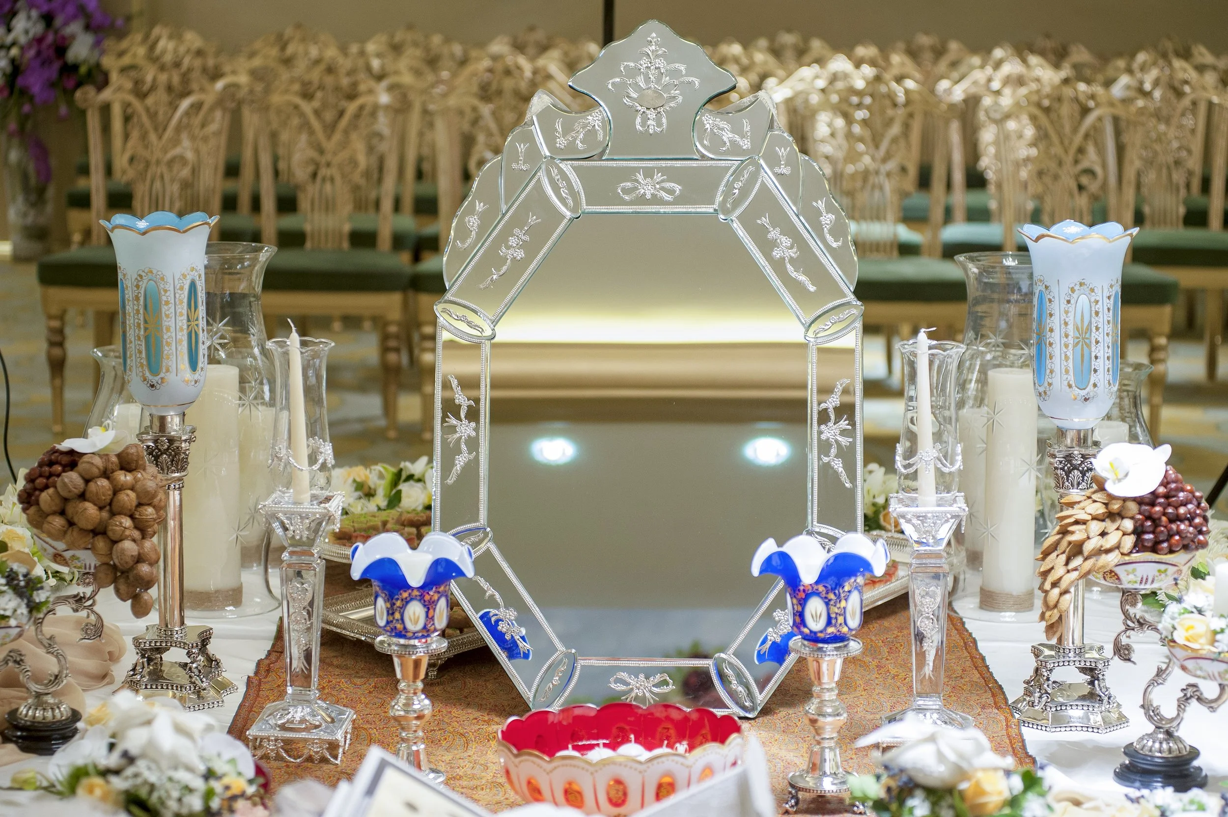 An elaborate, formal indoor table setting for a Persian wedding ceremony (Sofreh Aghd) featuring a large ornate mirror, silver candelabras with blue glass, nuts, candles, and white flowers.