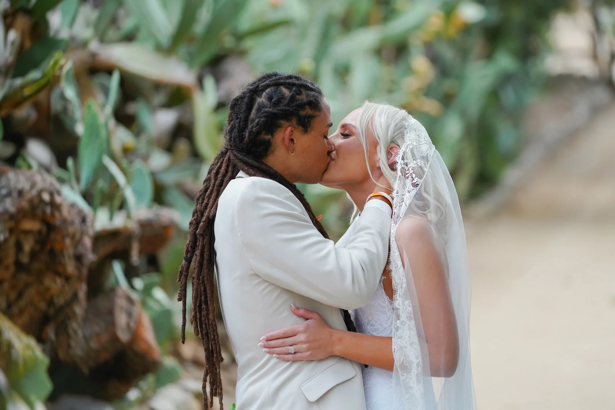 A photo of two brides sharing a tender kiss during their wedding ceremony, with one wearing a cream-colored suit and the other wearing a white dress with a lace-trimmed veil in Leo Garrillo Ranch Historic Park.